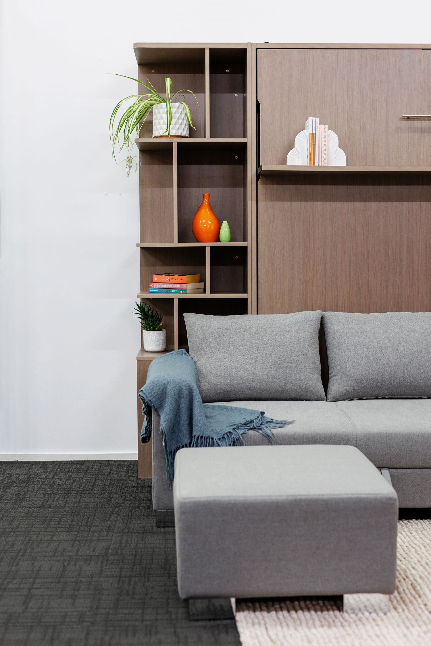 Modern living room interior with gray sectional sofa, ottoman and built-in shelving displaying decorative items.