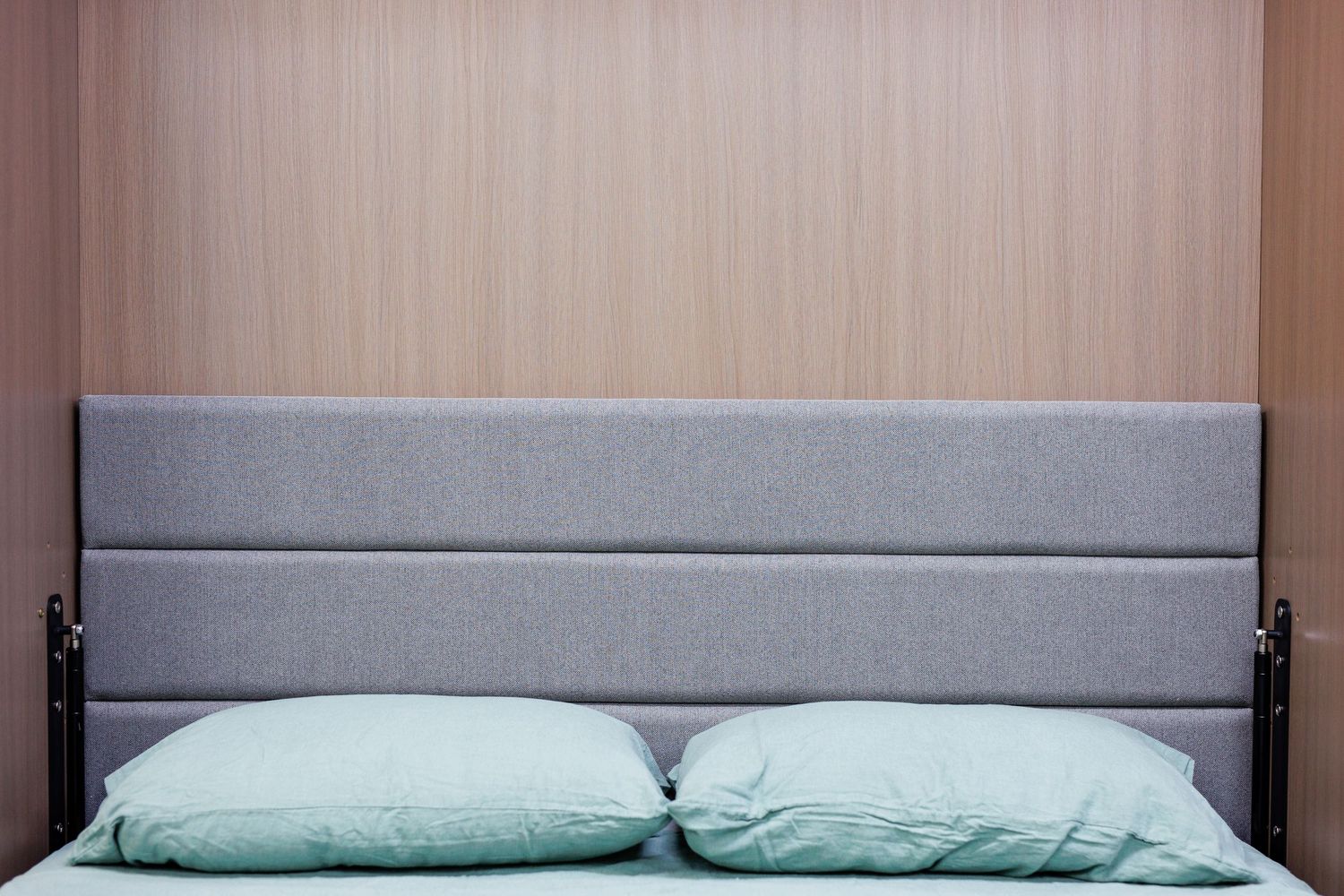Grey upholstered headboard with mint green pillows against light wood paneled wall.