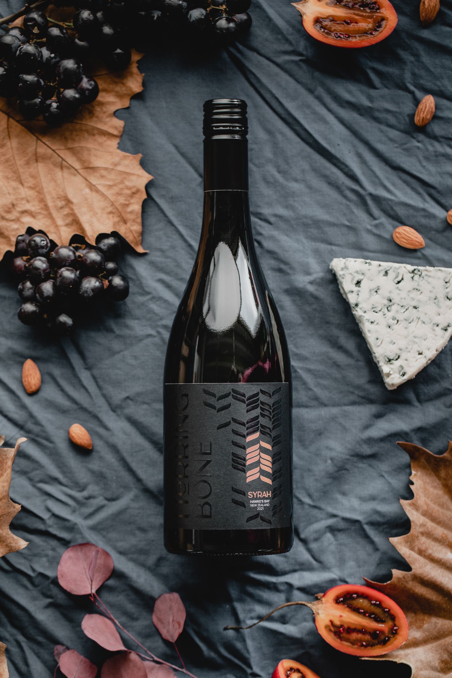 Black wine bottle surrounded by dark grapes, almonds, and fruit on a gray textile background.