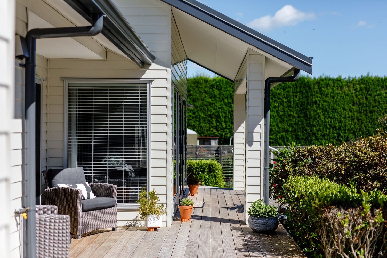 Cozy deck with wicker furniture and potted plants against white siding and green hedge.