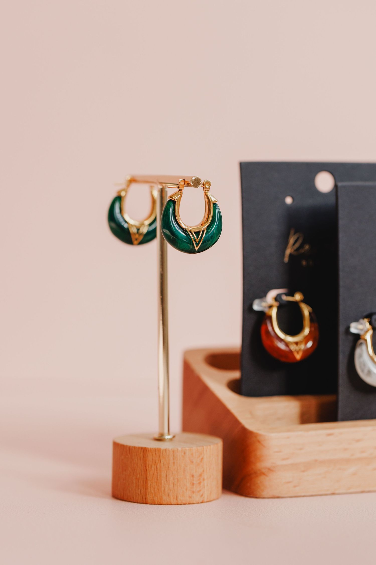 Elegant gold and green enamel jewelry displayed on wooden stands against a blush pink backdrop.