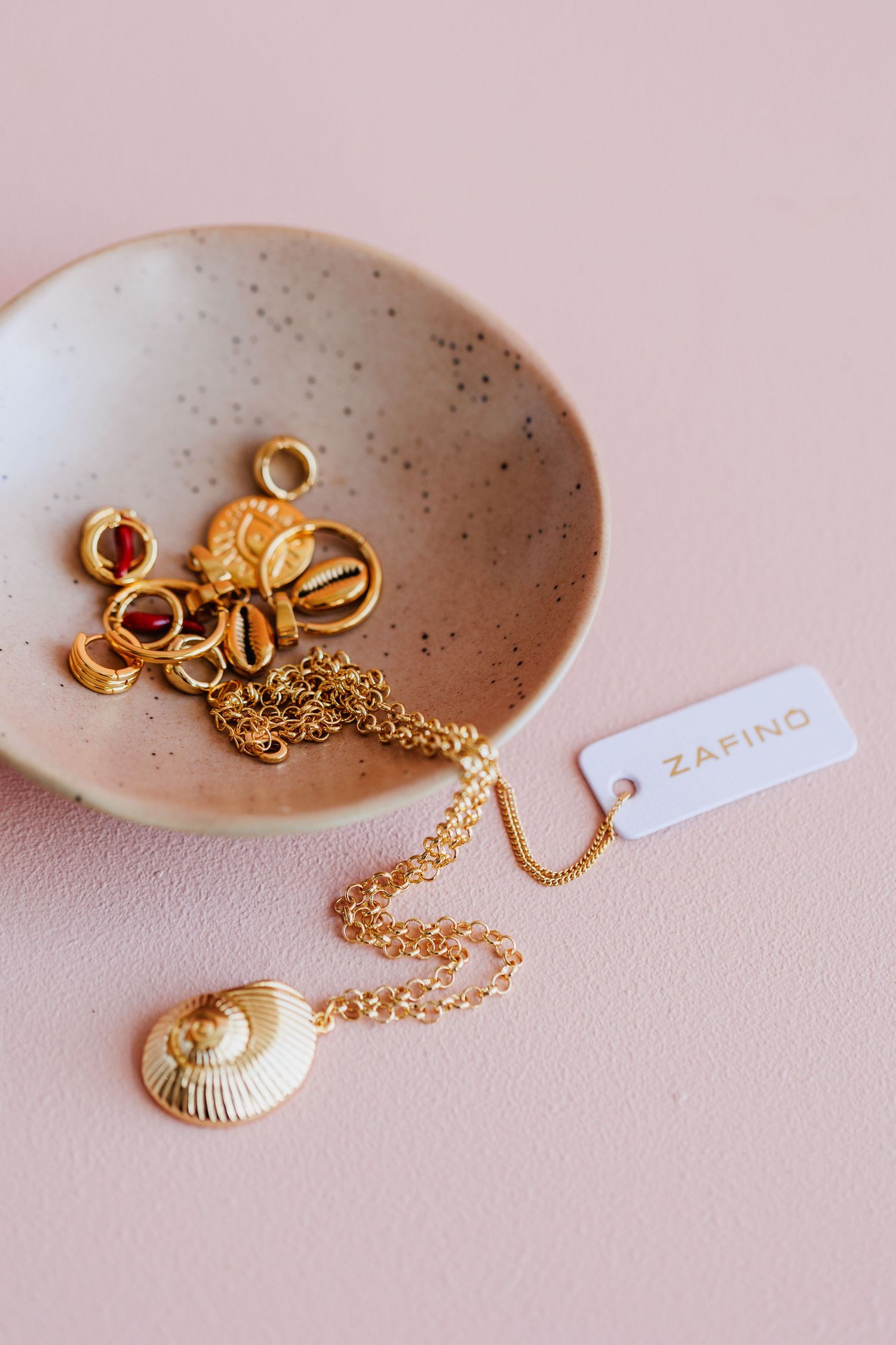 Gold chain necklace with shell pendant and charms displayed in a speckled ceramic dish.