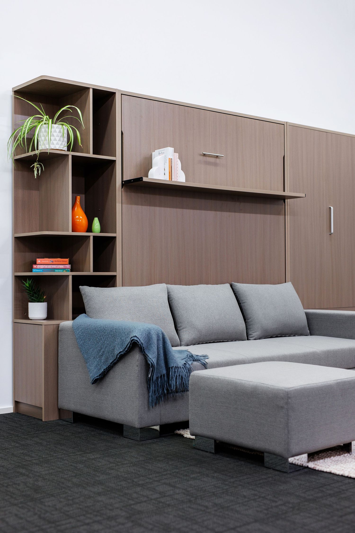 Modern murphy bed unit with grey sectional sofa and built-in shelving displaying plants and decor.