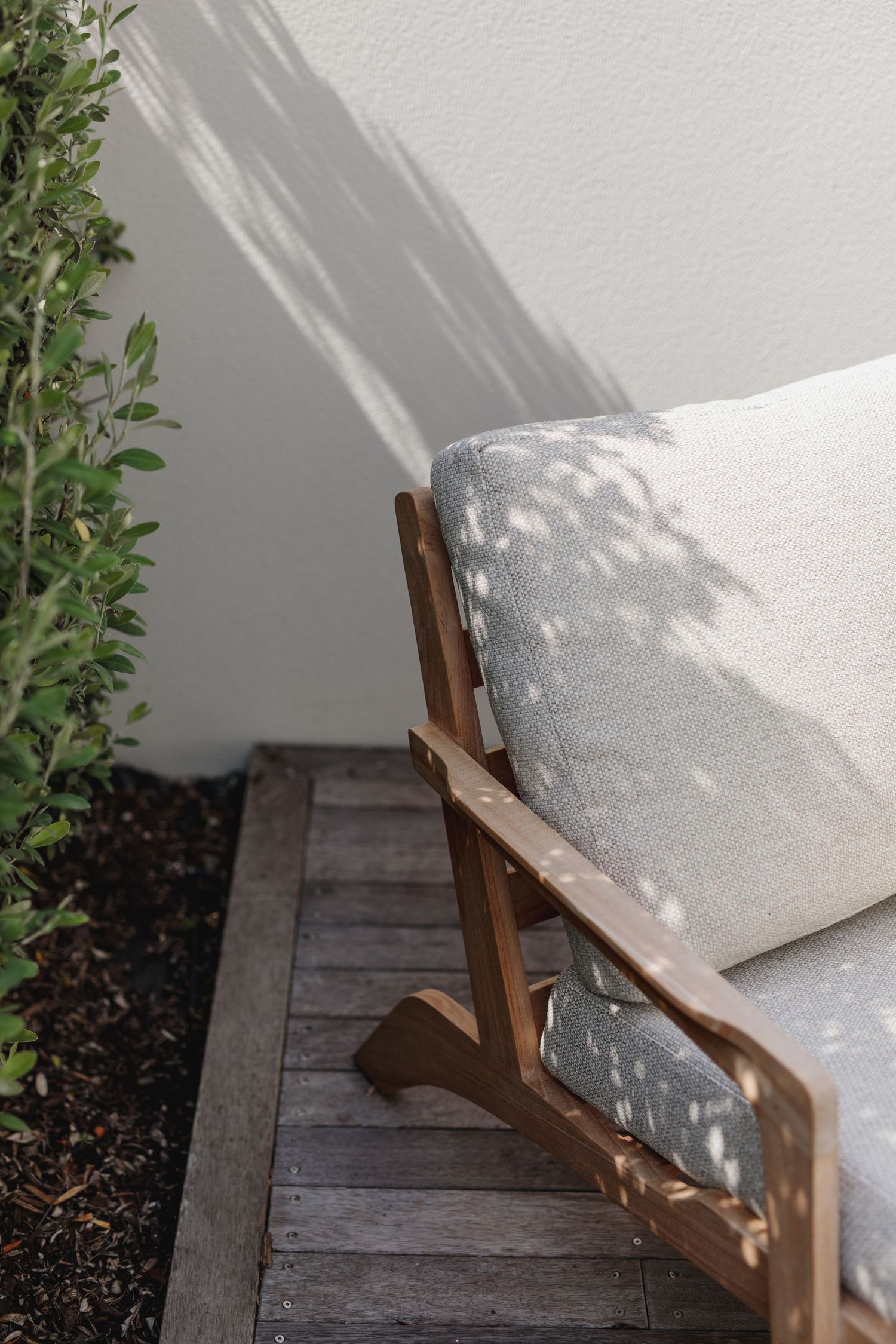 Wooden outdoor chair with white cushions casting dappled shadows on a deck.