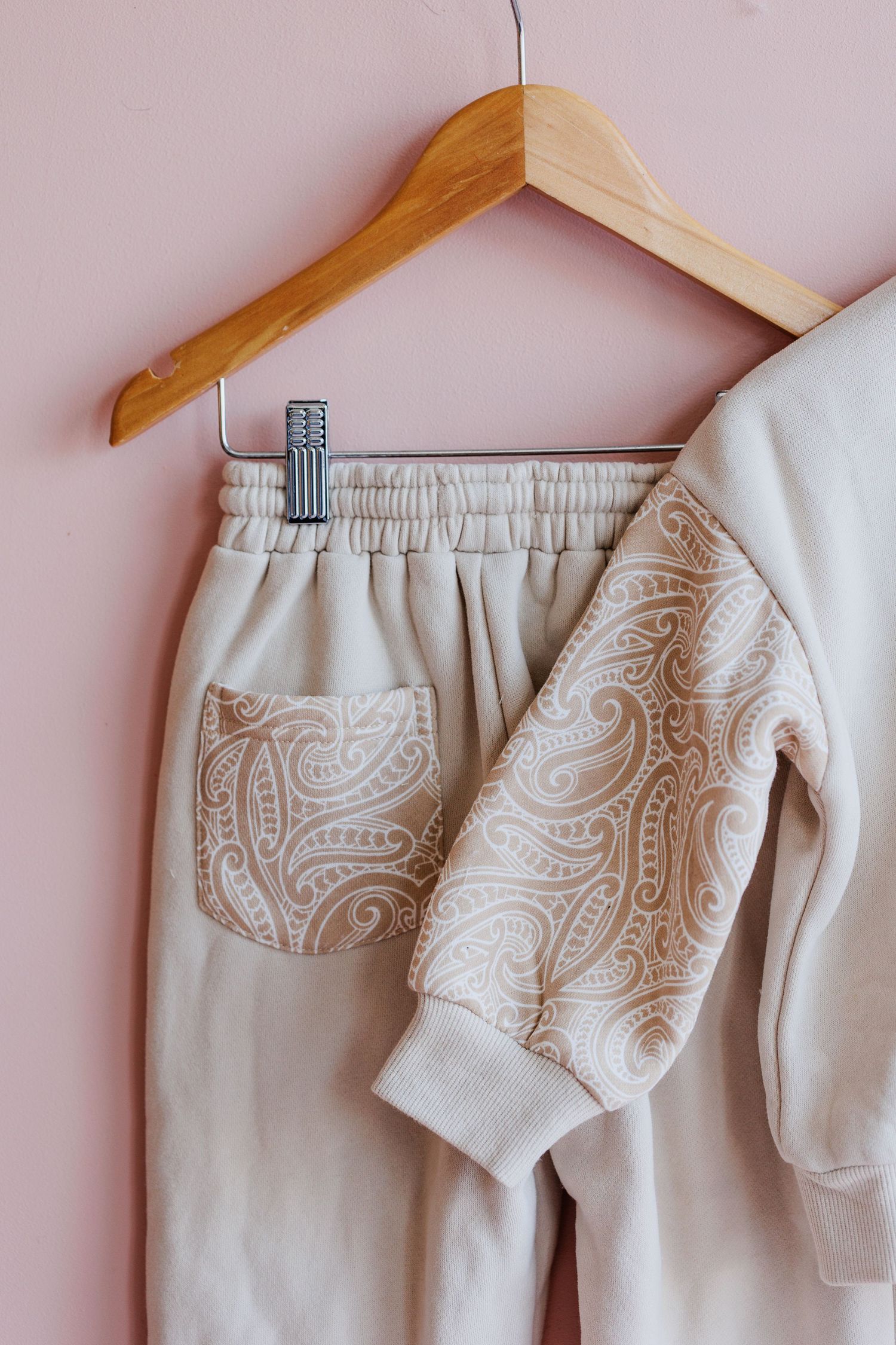 Beige sweatpants with paisley pattern hanging on wooden hanger against pink background.