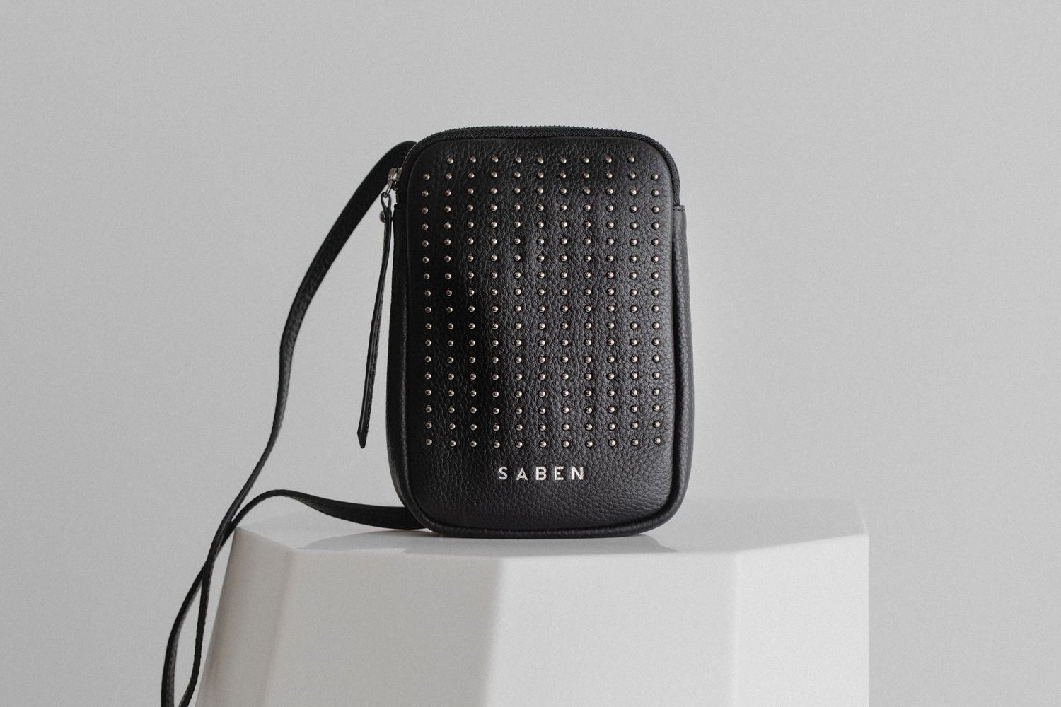 Black studded leather handbag displayed on white pedestal against gray background.