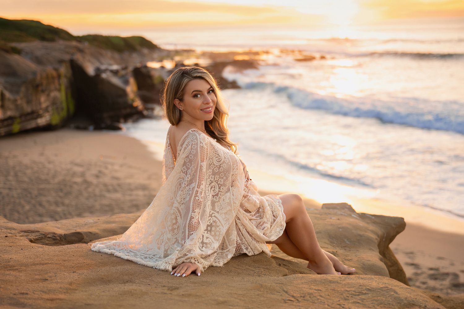 Smiling pregnant woman in cream lace dress sits on sandy beach rocks during warm golden sunset.