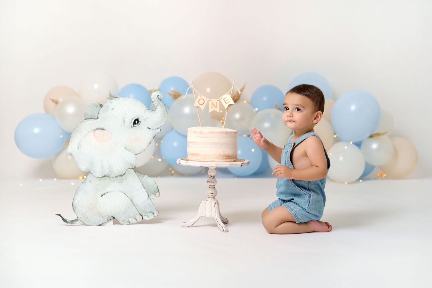 Baby boy in blue romper sits beside dinosaur toy and pink cake, surrounded by blue and beige balloons.