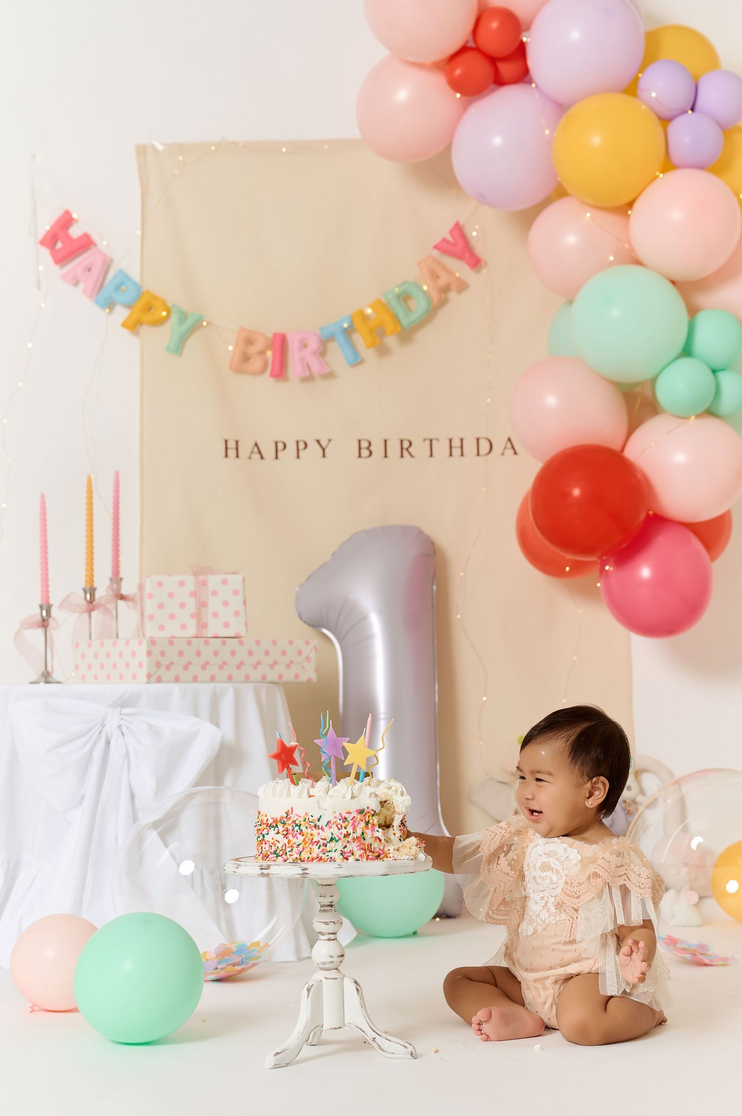 Baby girl celebrates first birthday with colorful balloons, cake, and Happy Birthday banner decorations.