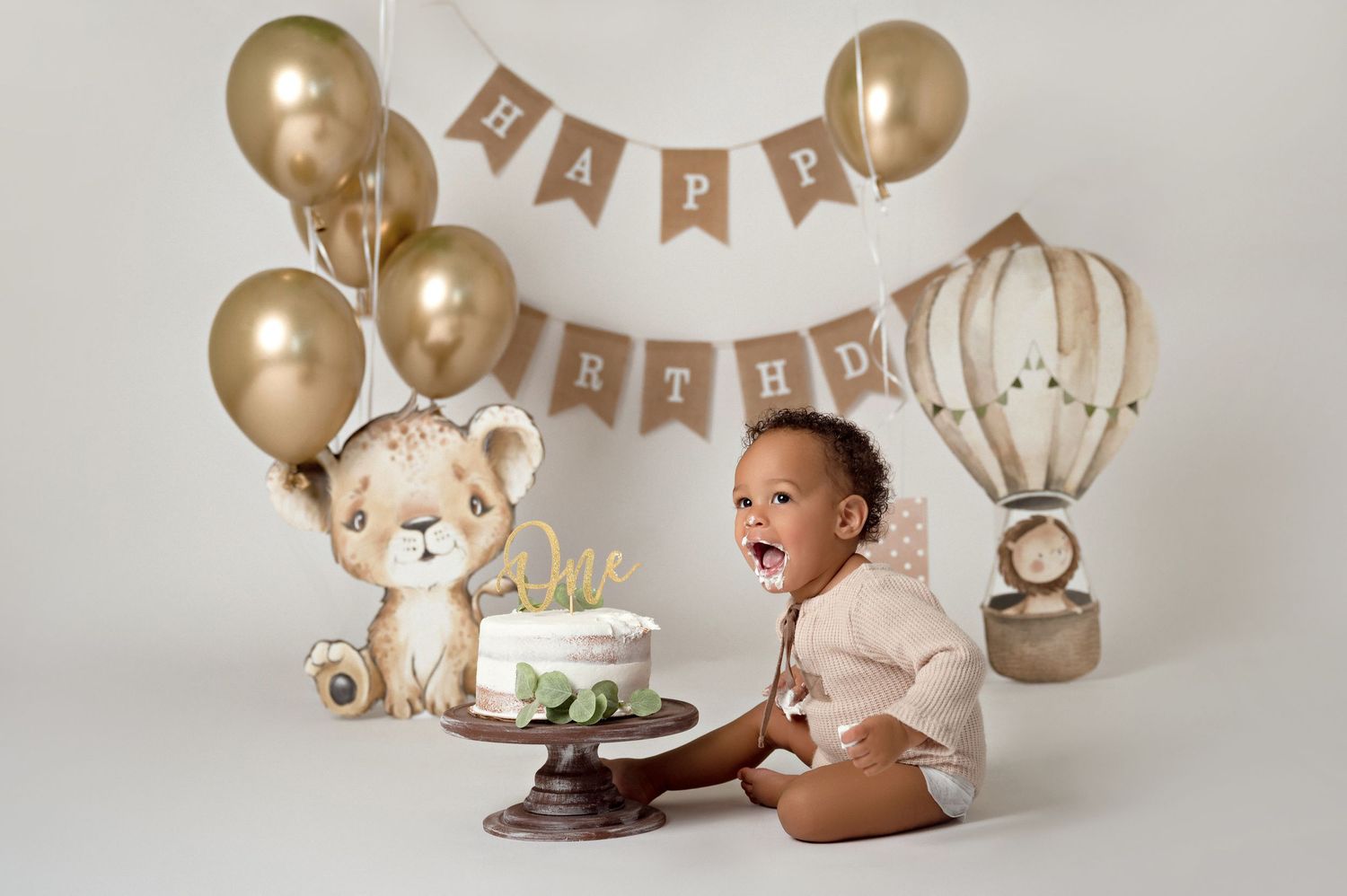 Baby's first birthday cake smash with gold balloons, lion cub decor, hot air balloon, and Happy Birthday banner.