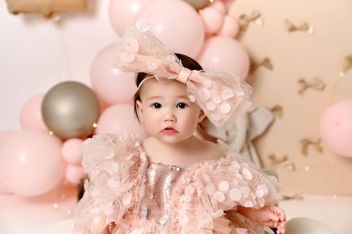 Baby girl in pink sequin dress with bow headband at first birthday cake smash with pink balloons.