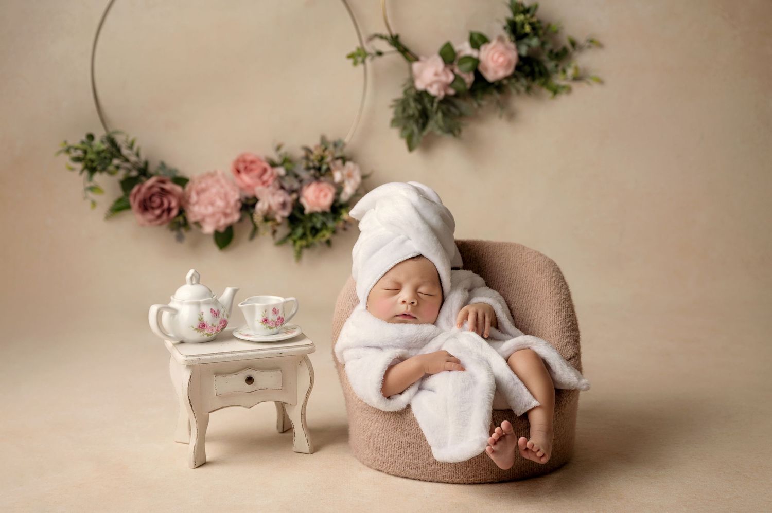 Newborn baby in white bathrobe and towel turban sitting in mini chair beside tiny tea set, floral hoop backdrop.