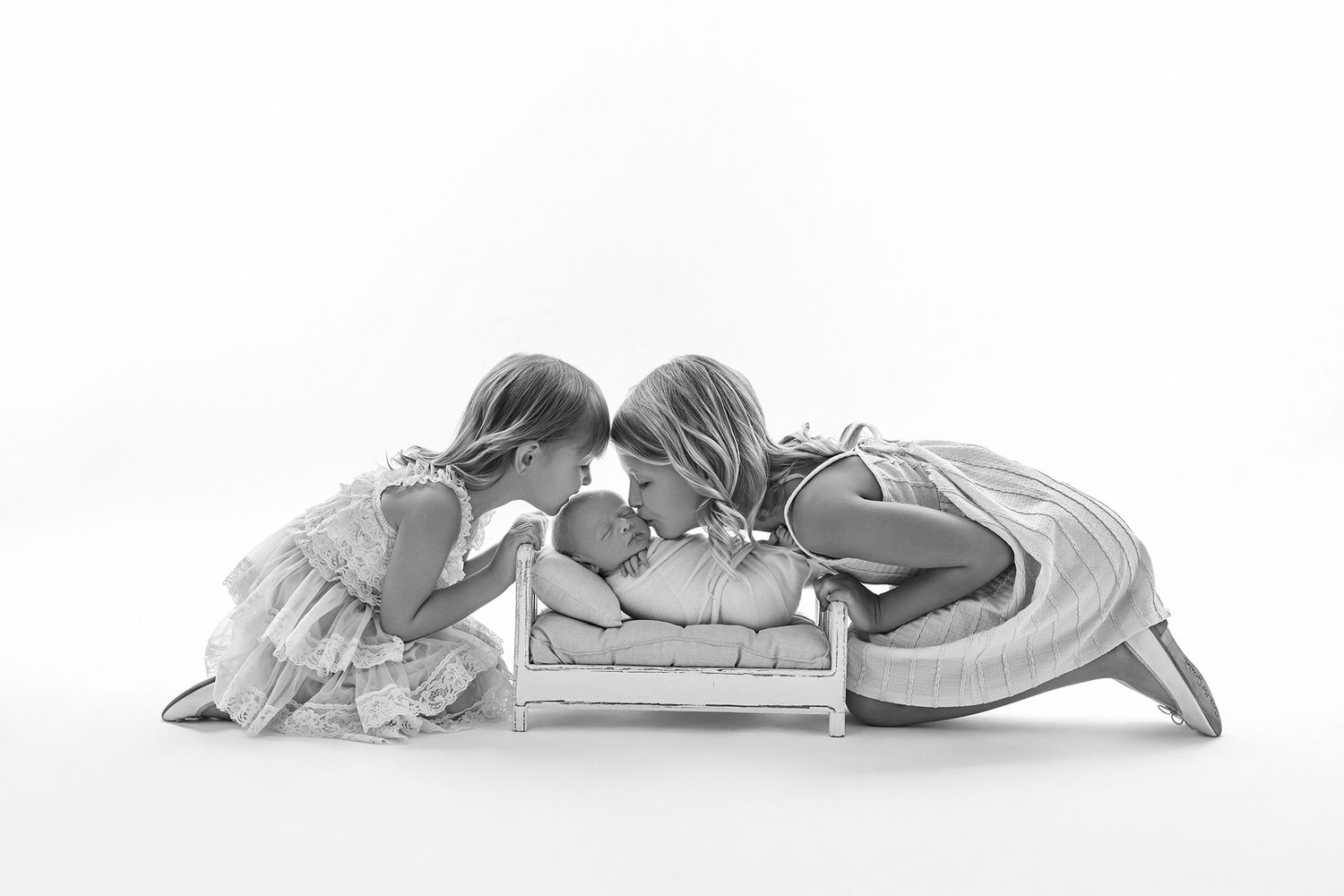 Black and white photo of two girls leaning in to kiss a newborn baby lying in a tiny wooden bed.