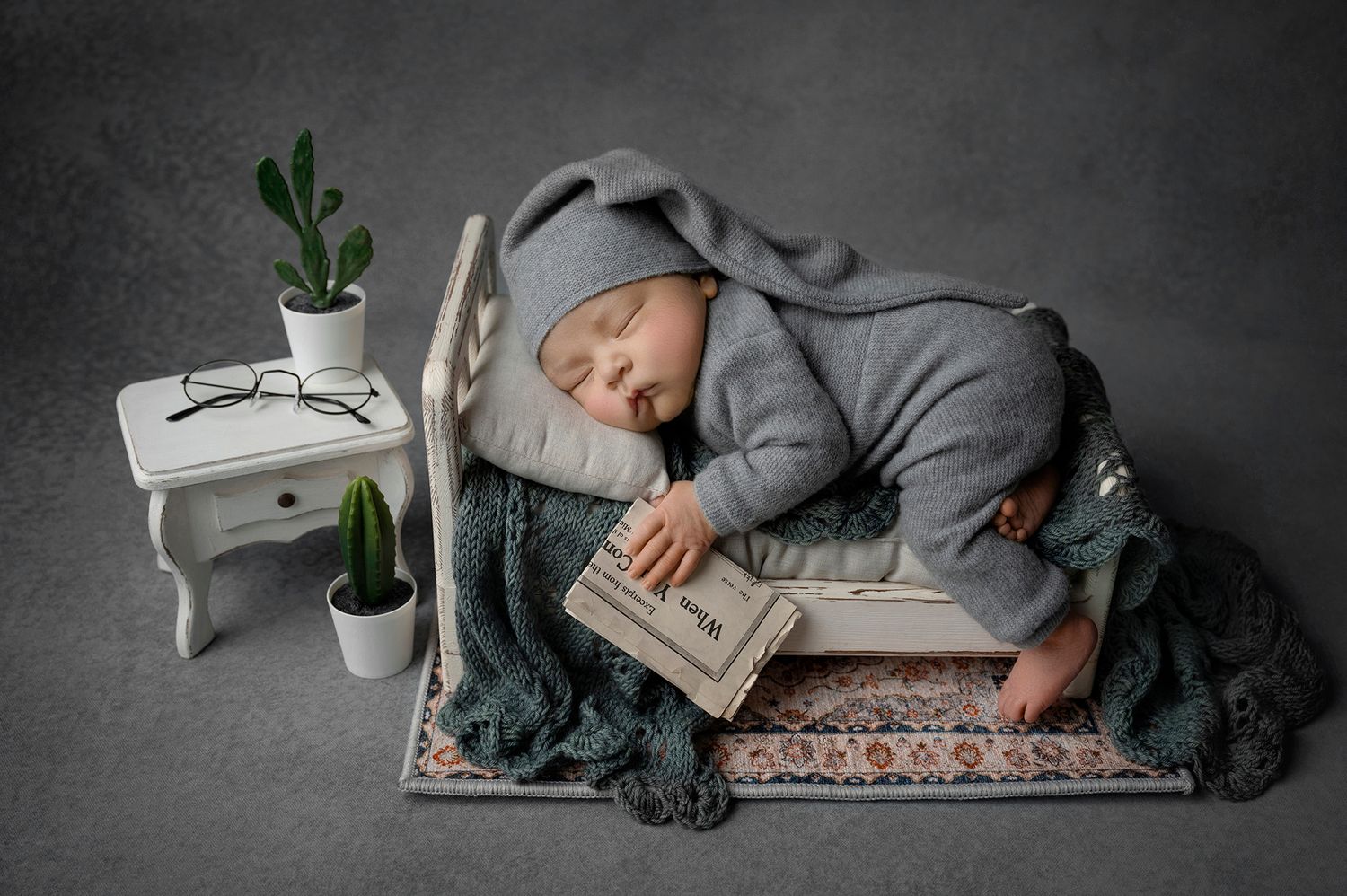 Sleeping newborn in gray bunny outfit posed on miniature bed with book and tiny plants.