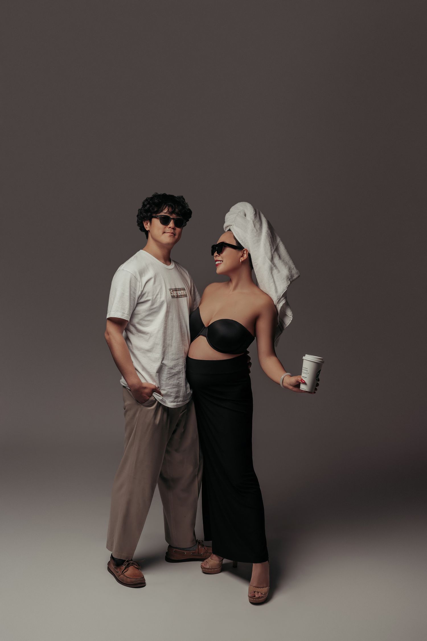 A stylish couple poses in sunglasses, man in white tee and beige pants, woman in black tube top with towel on head.