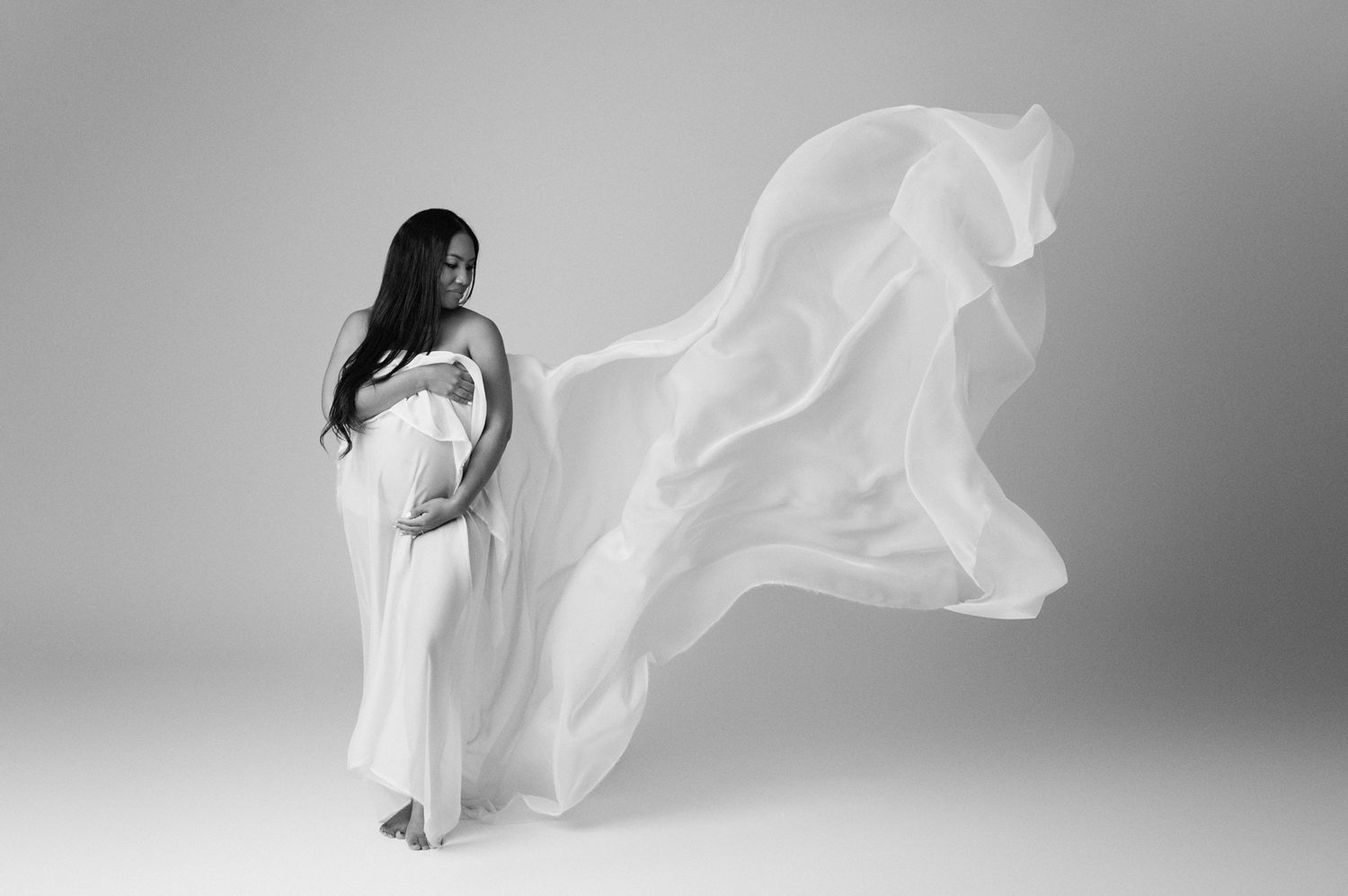 Black and white maternity photo of a woman in a flowing white gown with fabric billowing gracefully behind her.