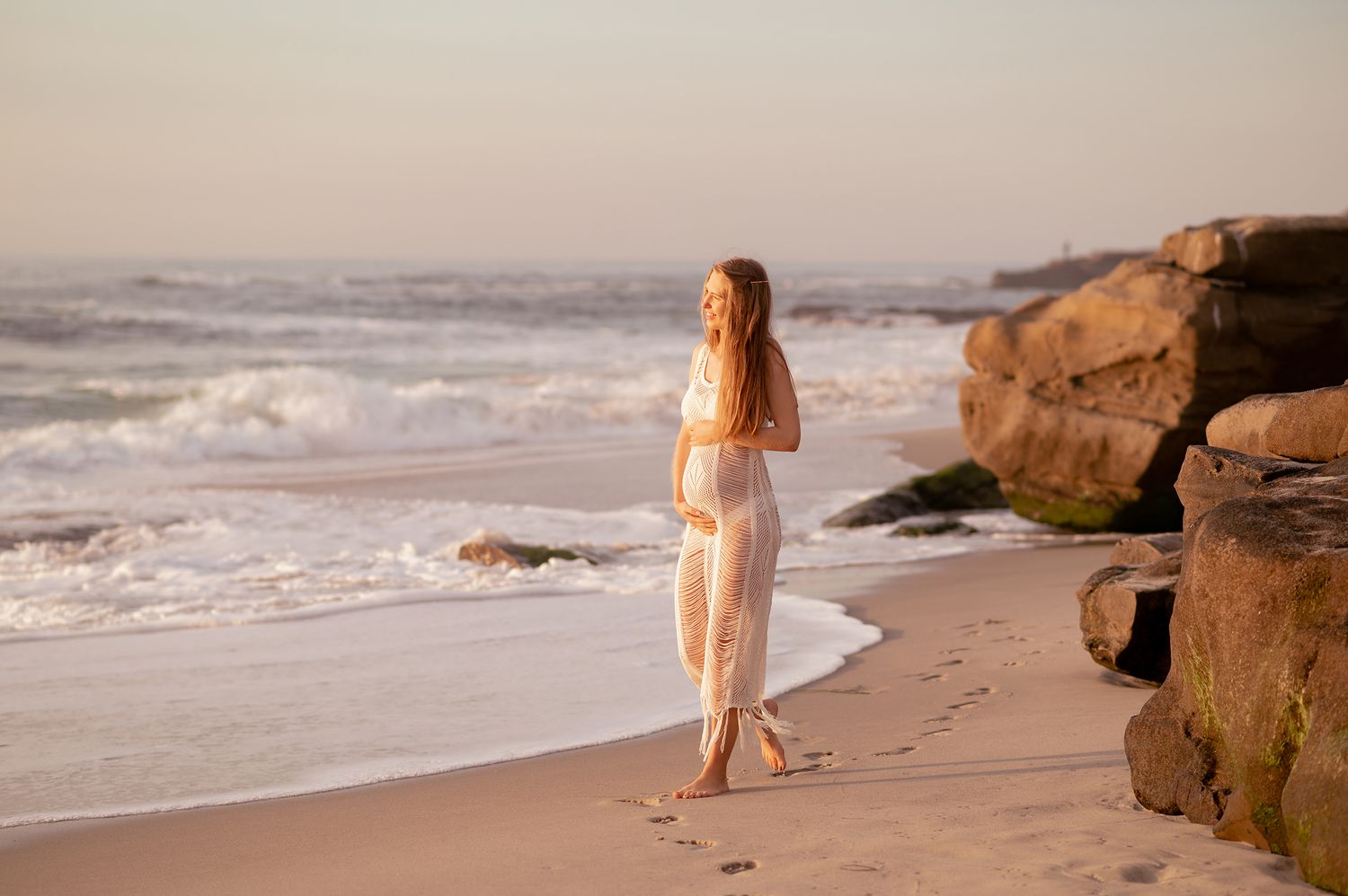 Pregnant woman in sheer white dress walks along sandy beach shoreline at soft pastel sunset.