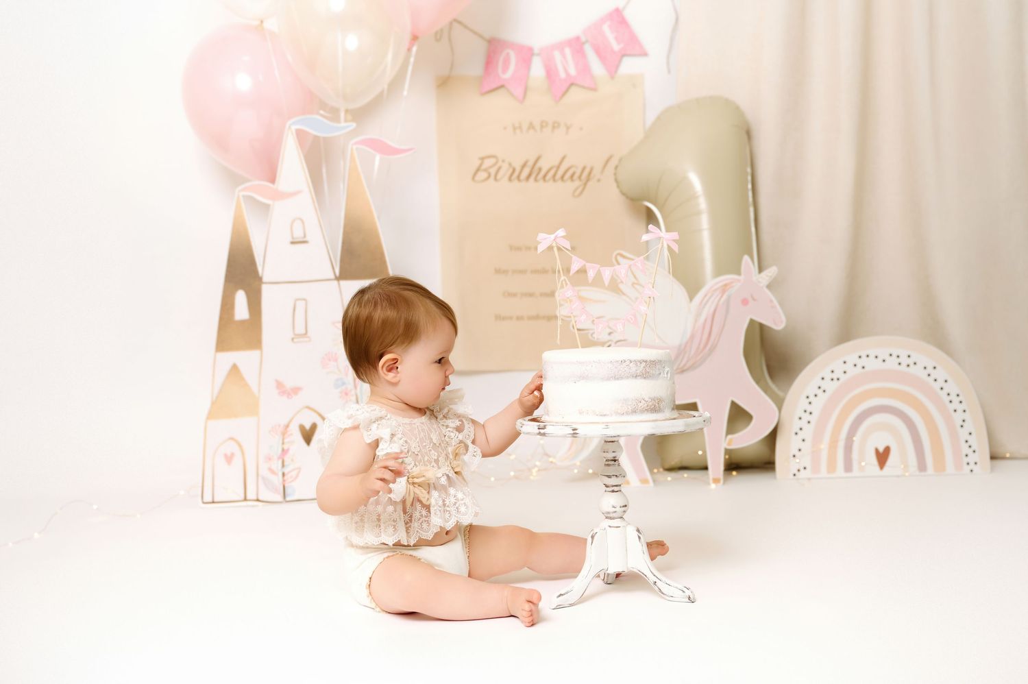 Baby girl sits at her first birthday cake smash setup with pink balloons, unicorn decor, and a Happy Birthday sign.