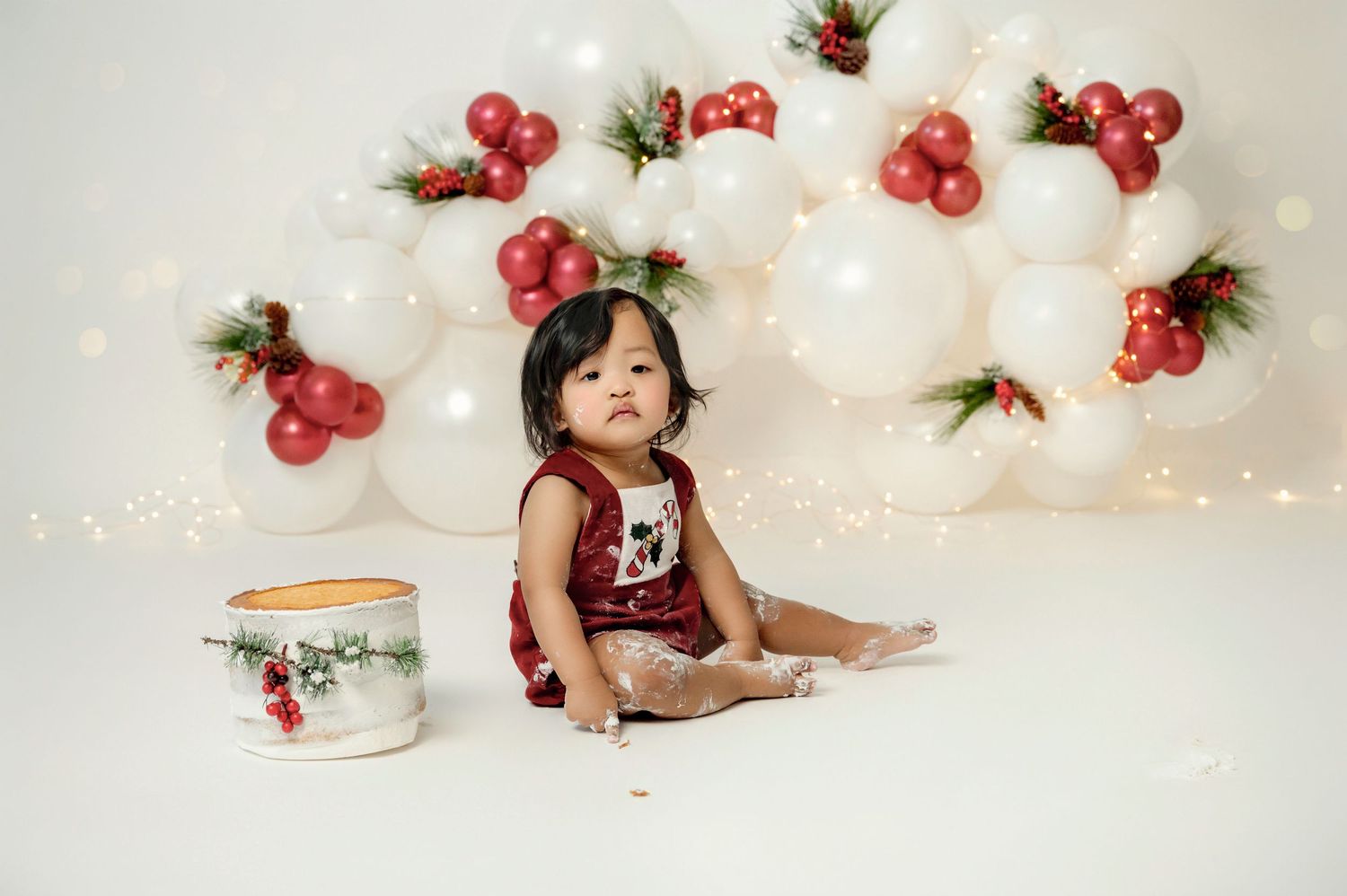 Baby girl in red romper sits by cake smash setup with white and red Christmas balloon backdrop.