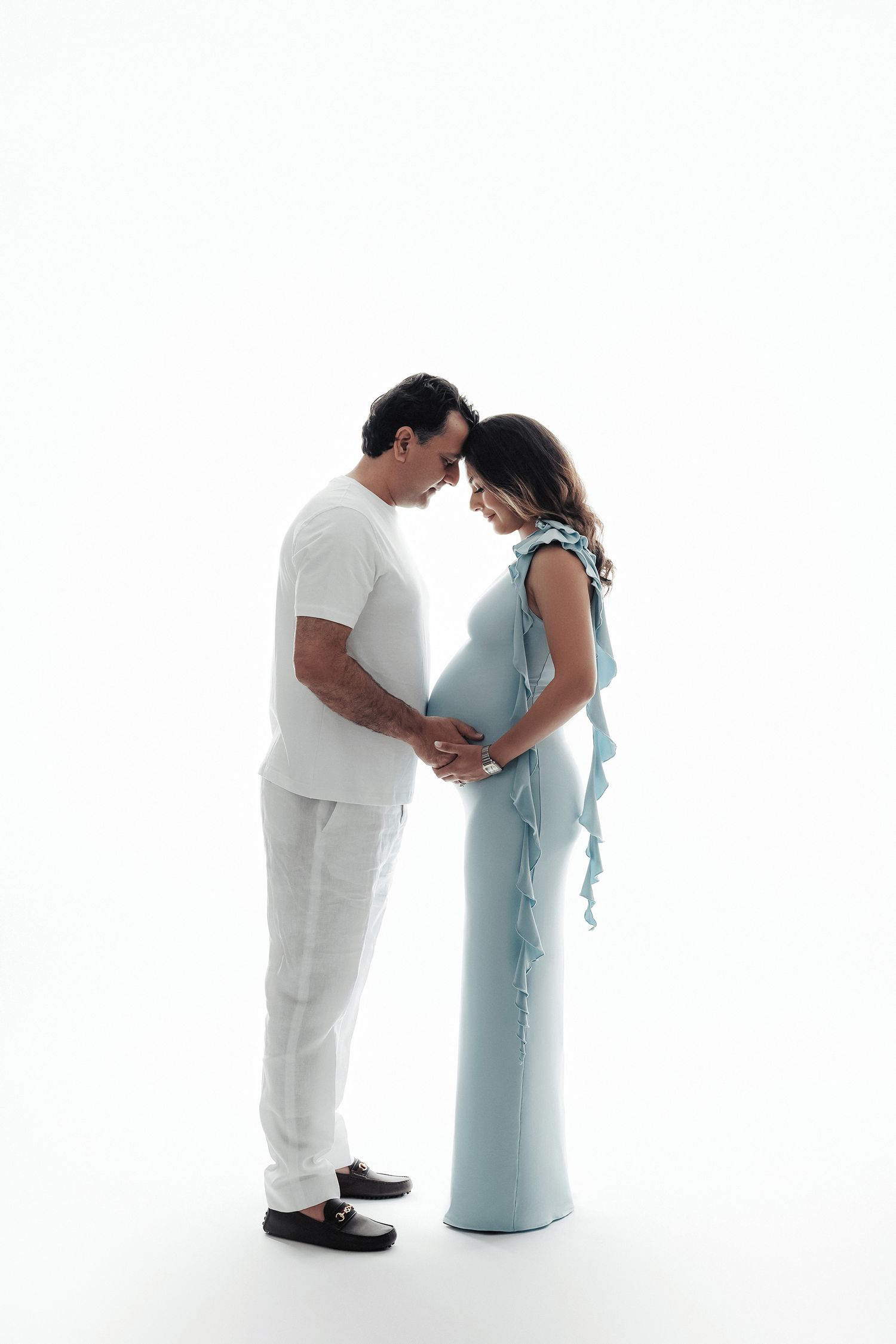 Expecting couple in white and blue outfits touch foreheads tenderly in bright white studio portrait.