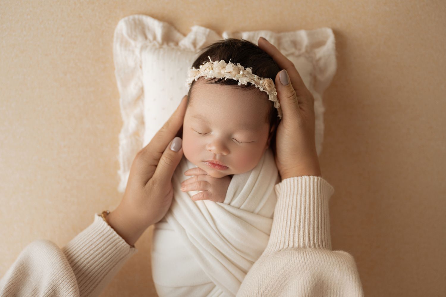 Newborn baby girl sleeping peacefully wrapped in white, wearing a floral headband, cradled by mother's hands.