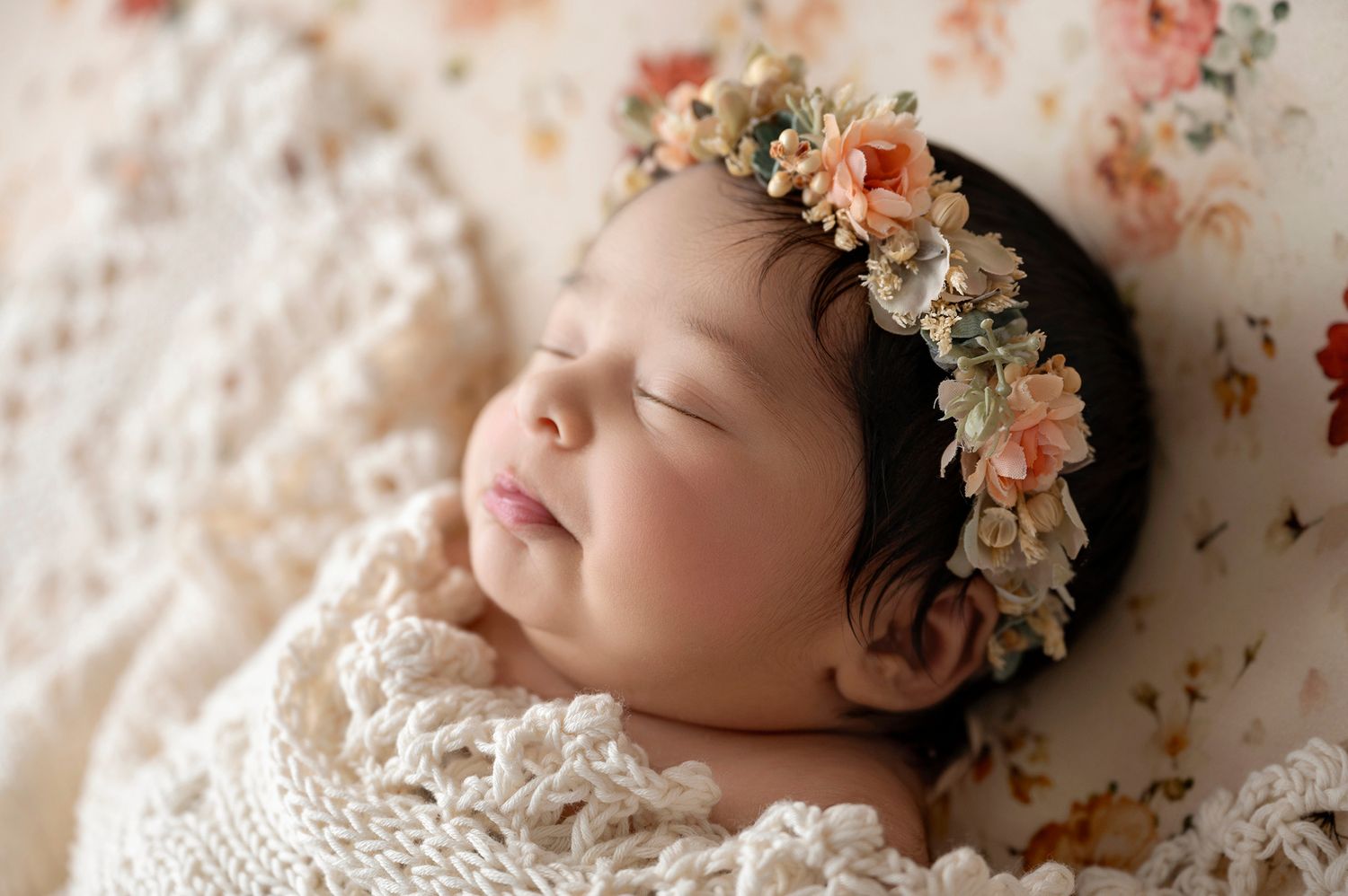 Sleeping newborn baby wrapped in cream knit blanket wearing a delicate floral headband on a floral background.
