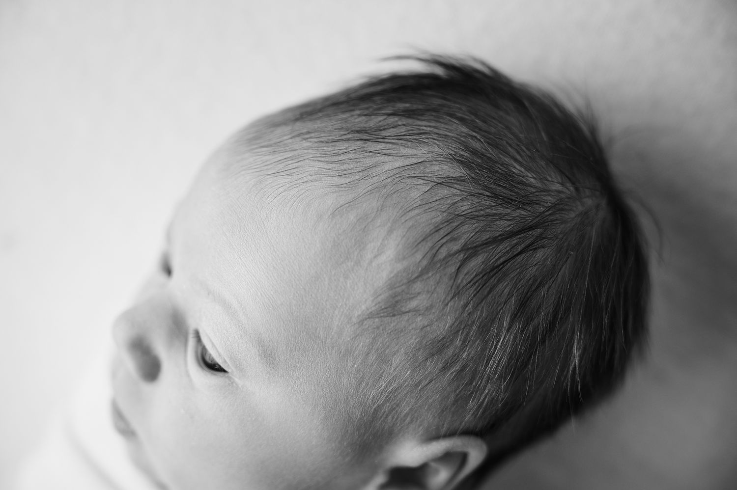 Black and white close-up photo of a newborn baby with fine dark hair lying peacefully on a soft white surface.