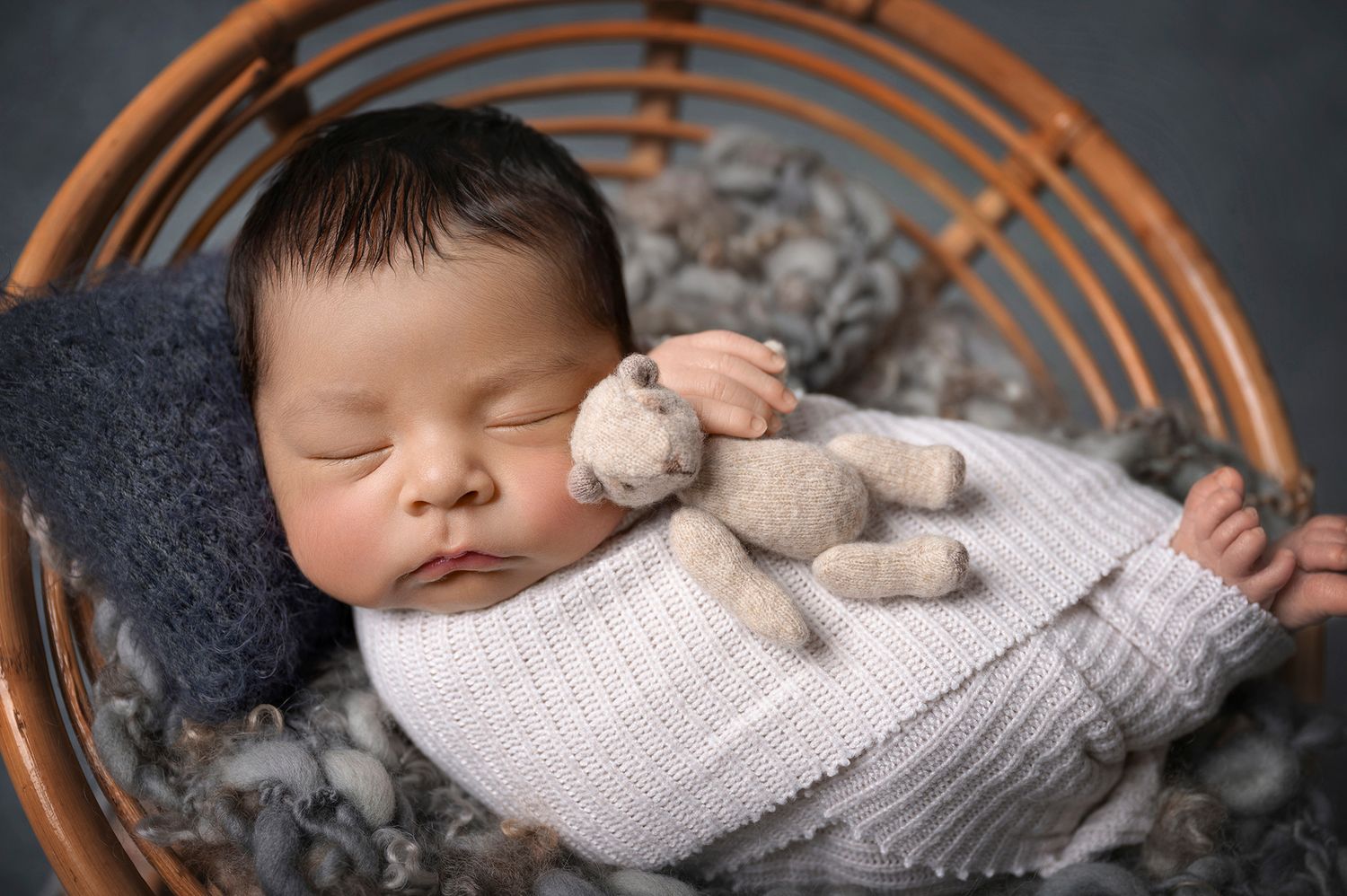 Newborn baby sleeping in wicker chair wrapped in white knit blanket holding a small teddy bear.