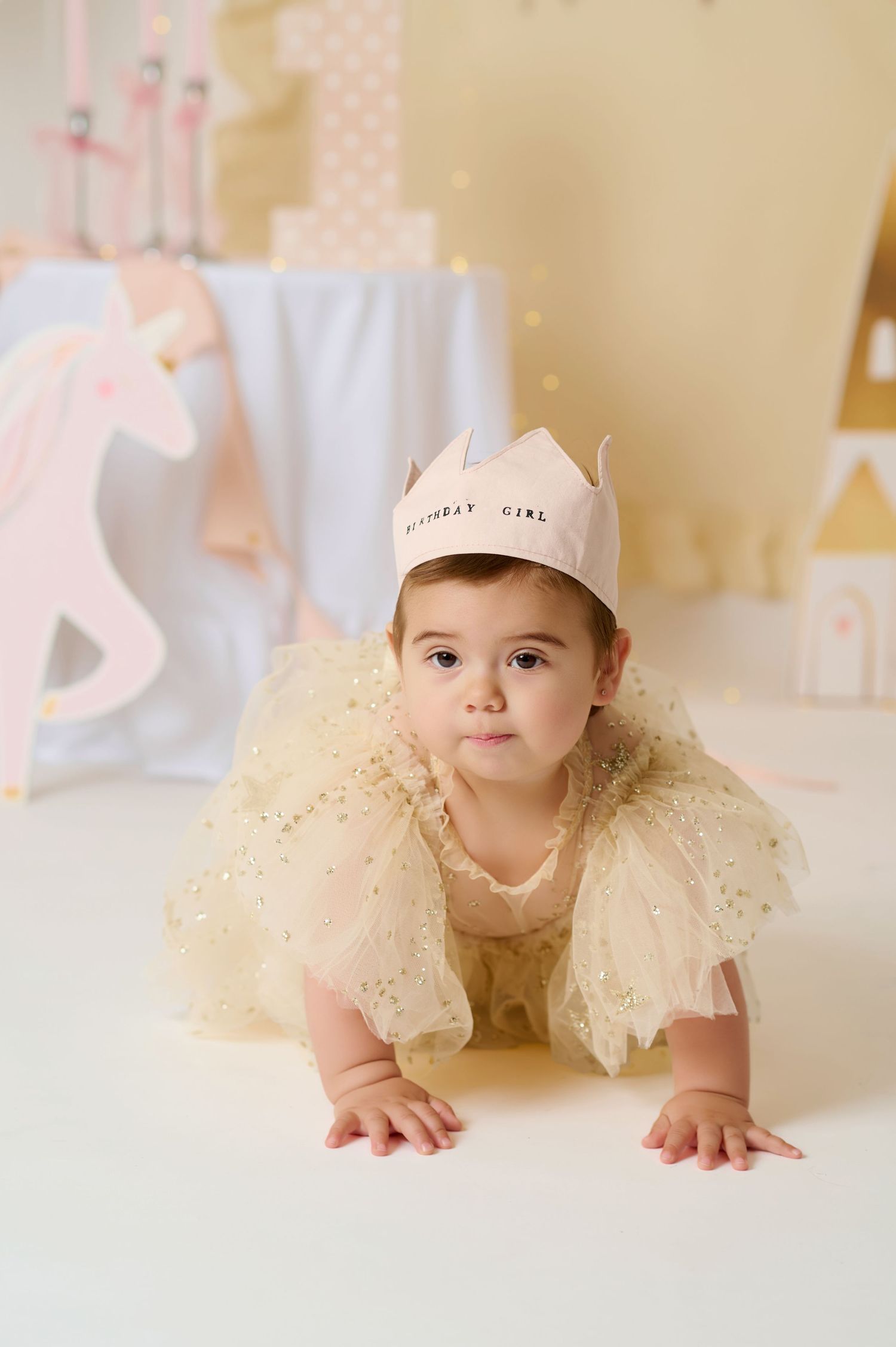 A baby girl in a yellow tulle dress and birthday crown crawls on a white floor with unicorn decorations behind her.