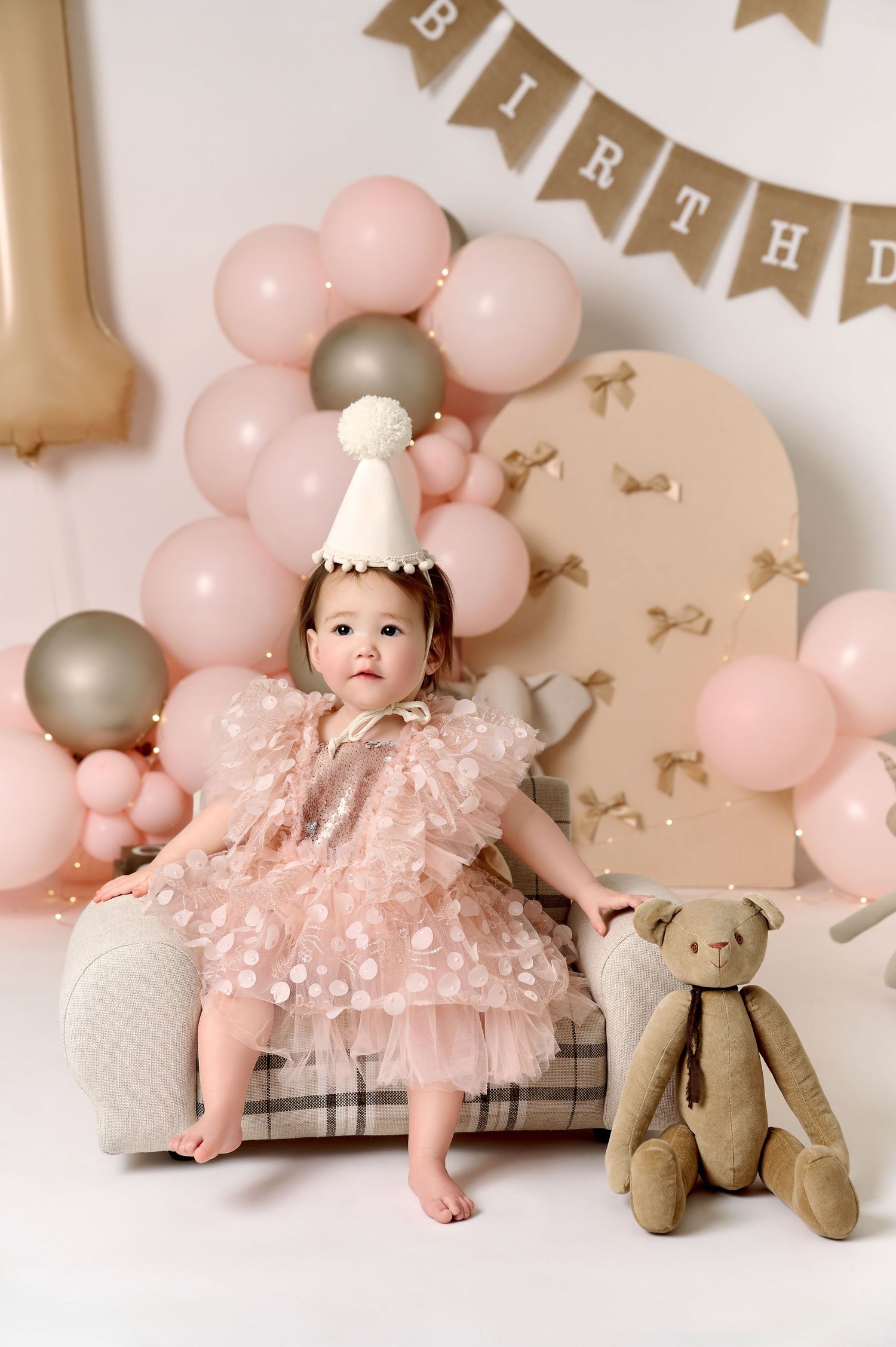 Baby girl in pink tutu dress and party hat sits beside teddy bear at a first birthday photo shoot.