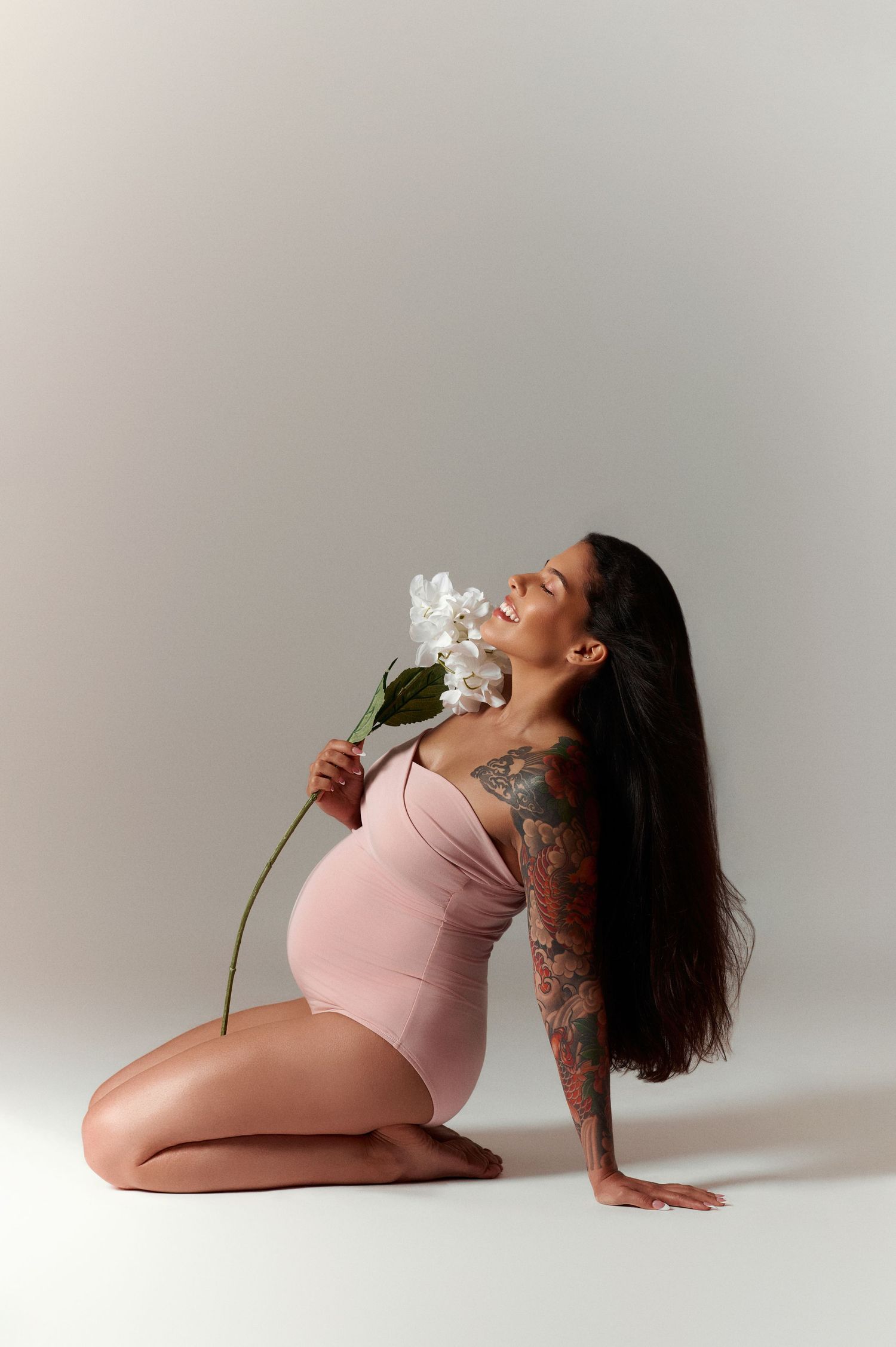 Pregnant woman with tattoos in pink bodysuit sitting on floor holding white flowers, smiling with long dark hair.