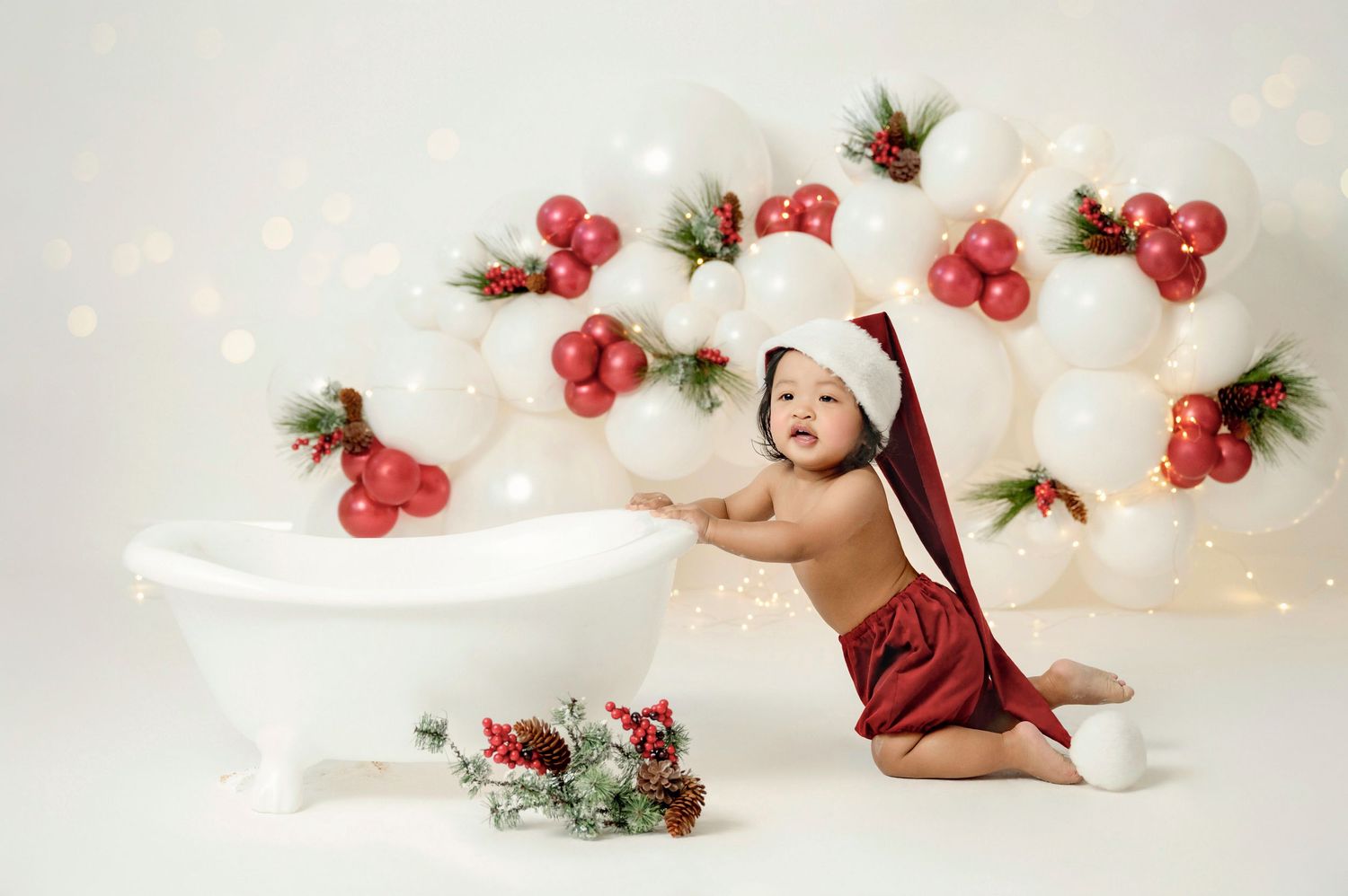 Baby in Santa hat poses by mini bathtub with festive red and white balloon arch and holiday decorations.