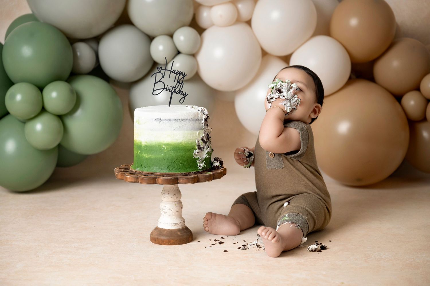 Baby boy in olive romper smashing first birthday cake surrounded by sage green and tan balloon backdrop.