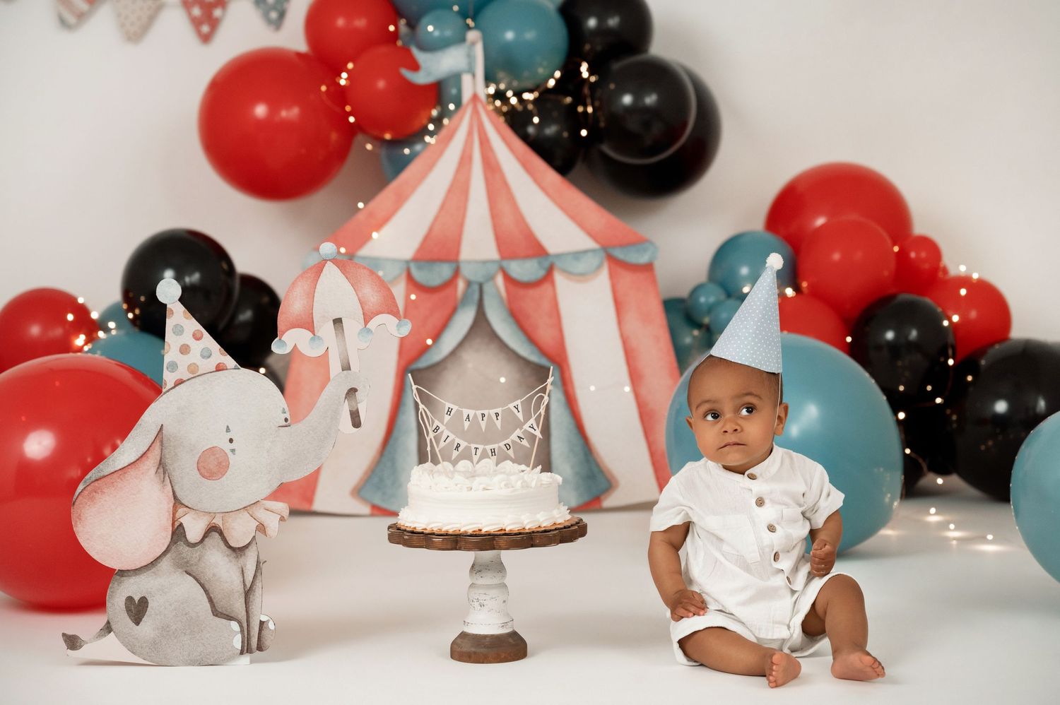 Baby's first birthday circus cake smash with red, black, and teal balloons, elephant decor, and striped tent backdrop.