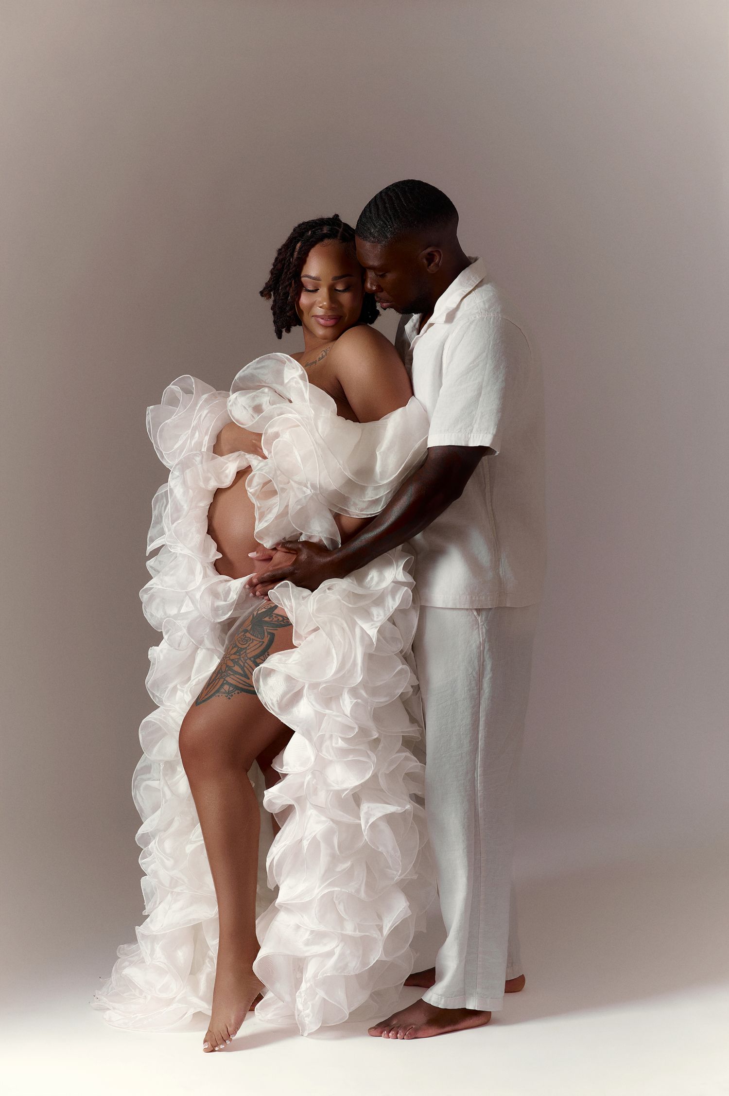 Pregnant Black couple in white outfits pose tenderly in studio, man embracing woman in ruffled maternity gown.