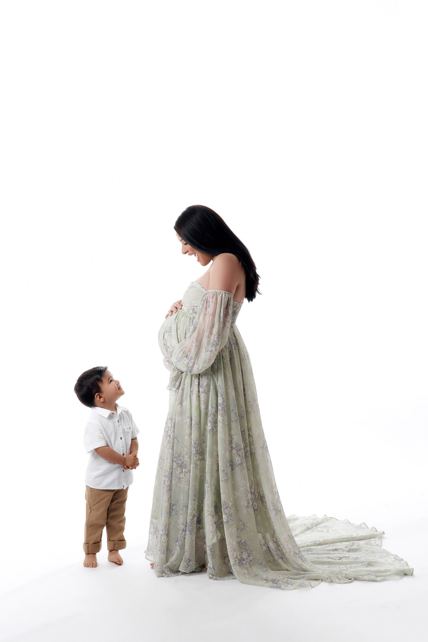 Pregnant woman in floral green gown with young son looking up at her baby bump in white studio.