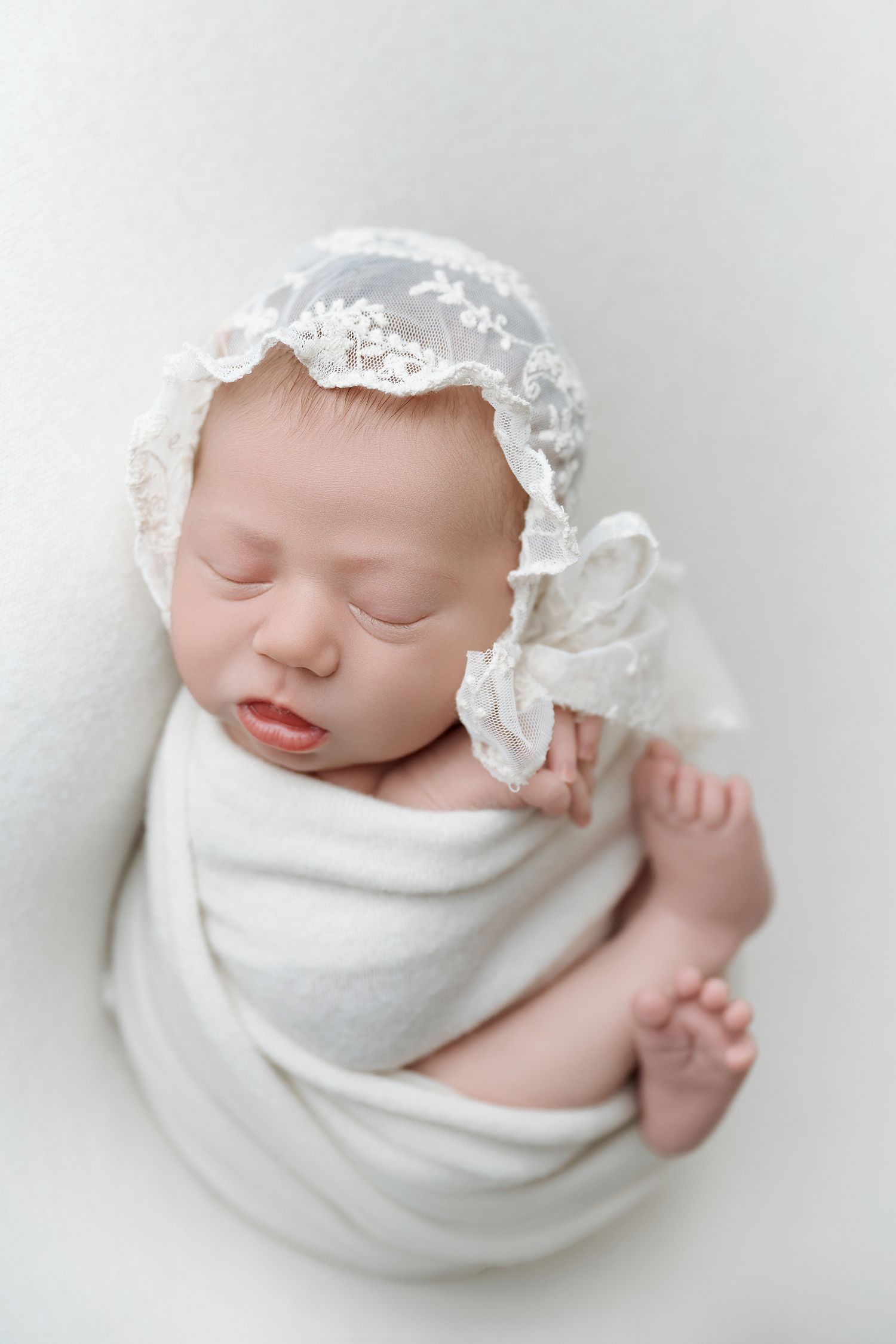 Sleeping newborn baby girl wrapped in white swaddle wearing delicate lace bonnet on white background.