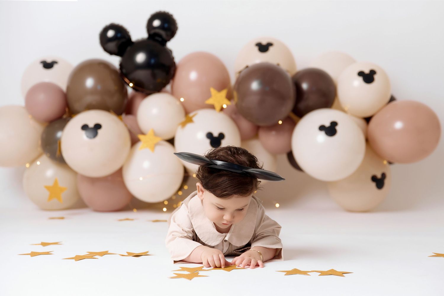 Baby in Mickey Mouse hat crawls among gold stars, surrounded by beige and black Mickey-themed balloons on white background.