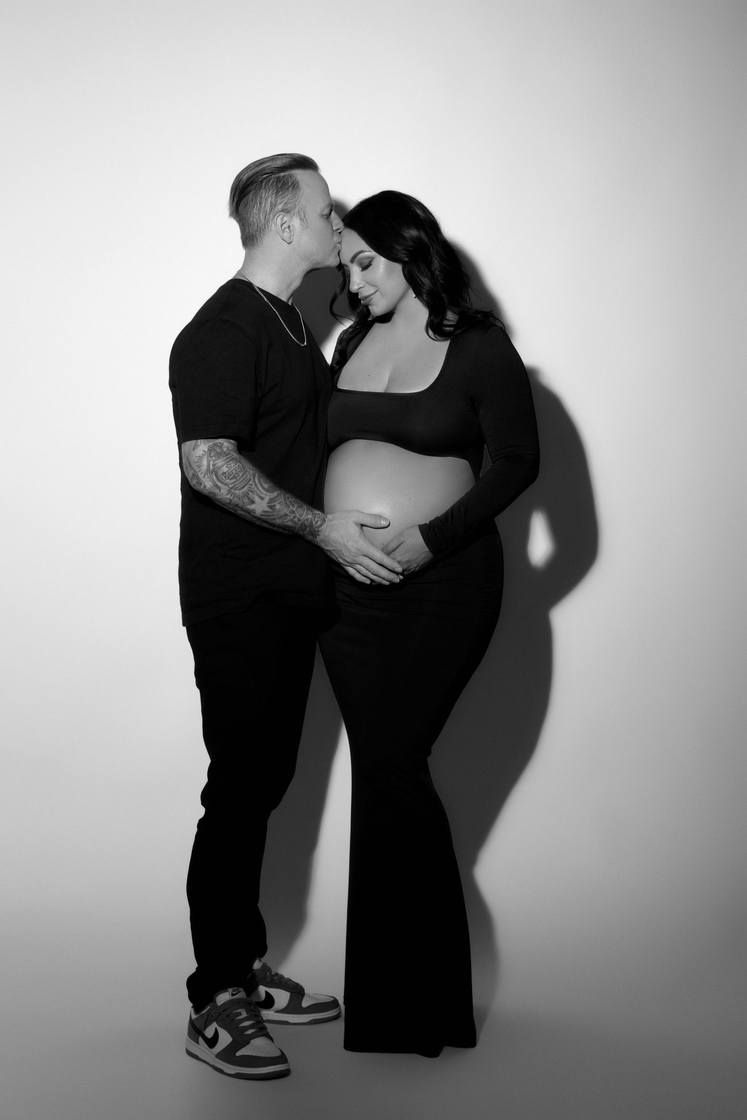 Black and white maternity photo of a couple in black outfits, man kissing woman's head while holding her baby bump.