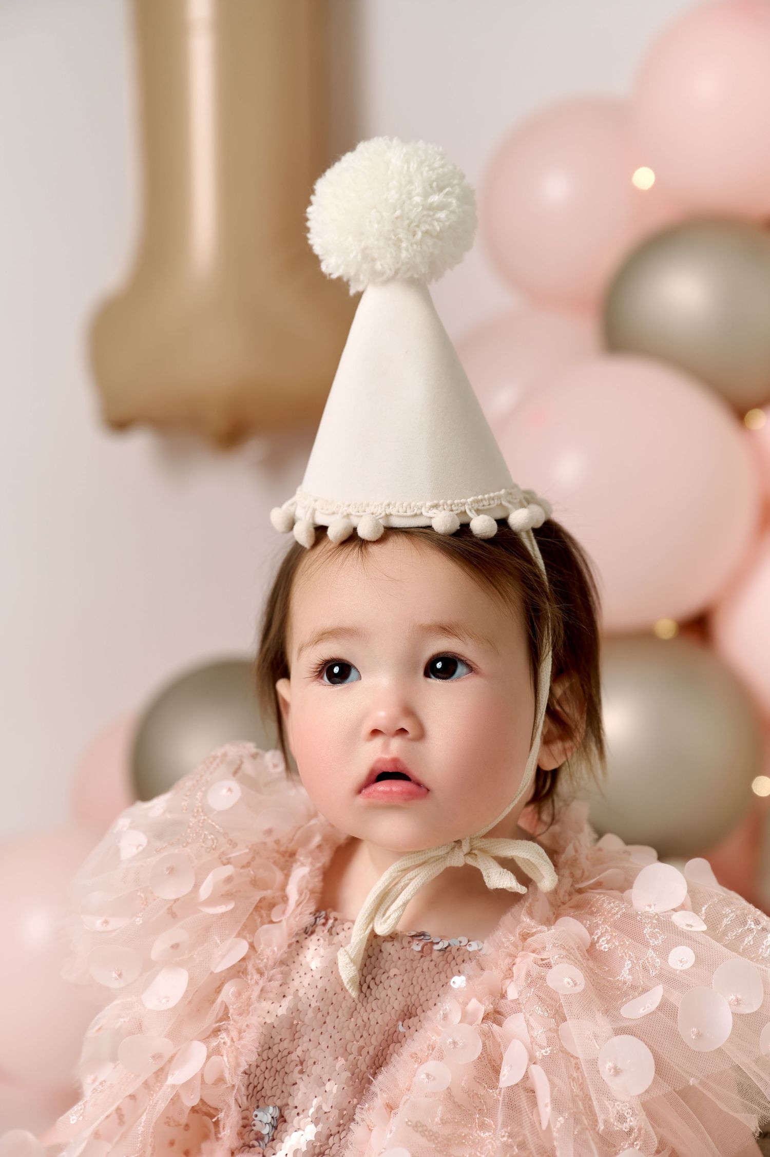 Adorable baby girl in pink sequin dress and white pom-pom party hat celebrates first birthday with balloons.