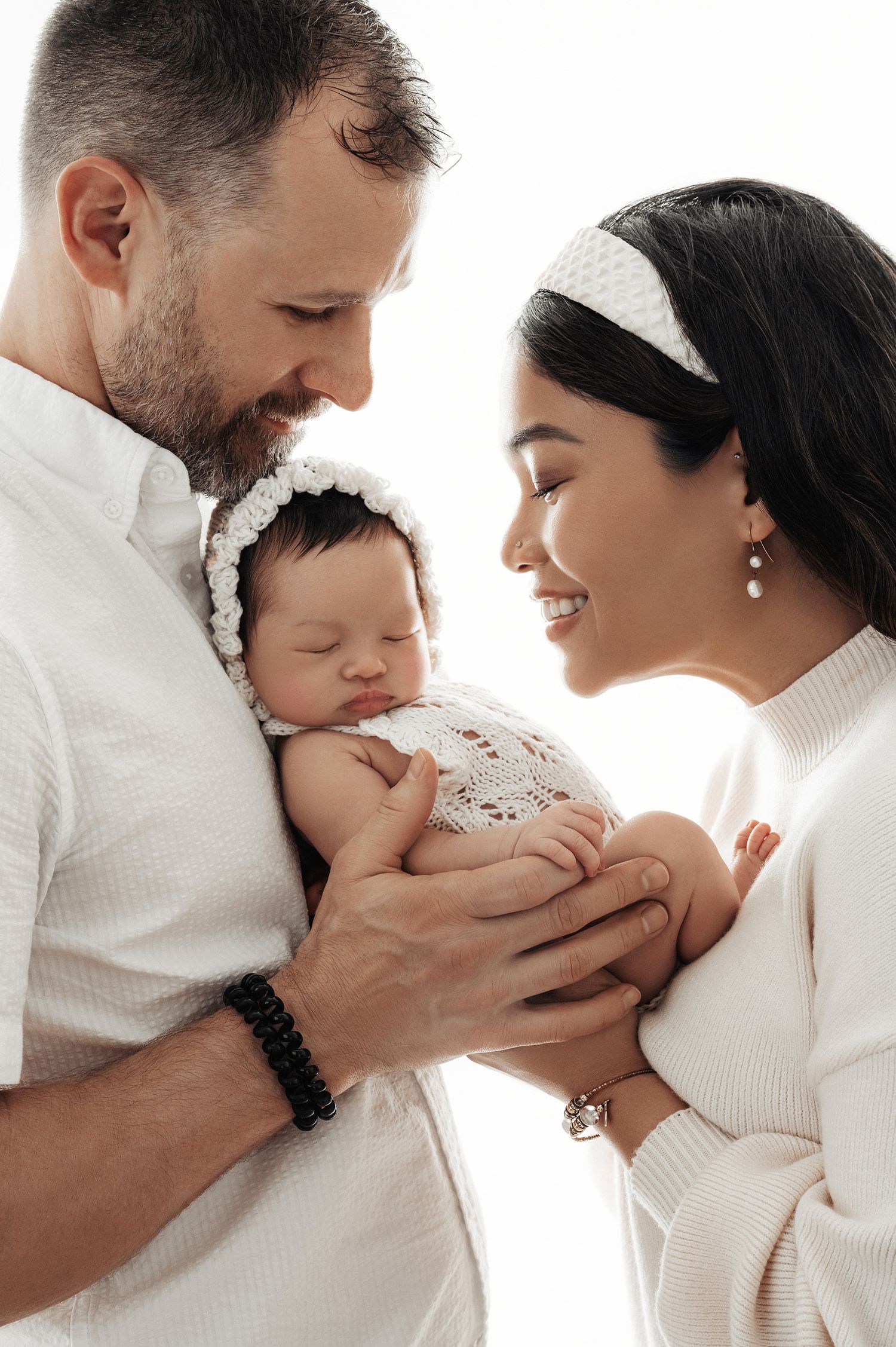 A couple in white clothing tenderly holds their sleeping newborn baby, dressed in a white crochet outfit.