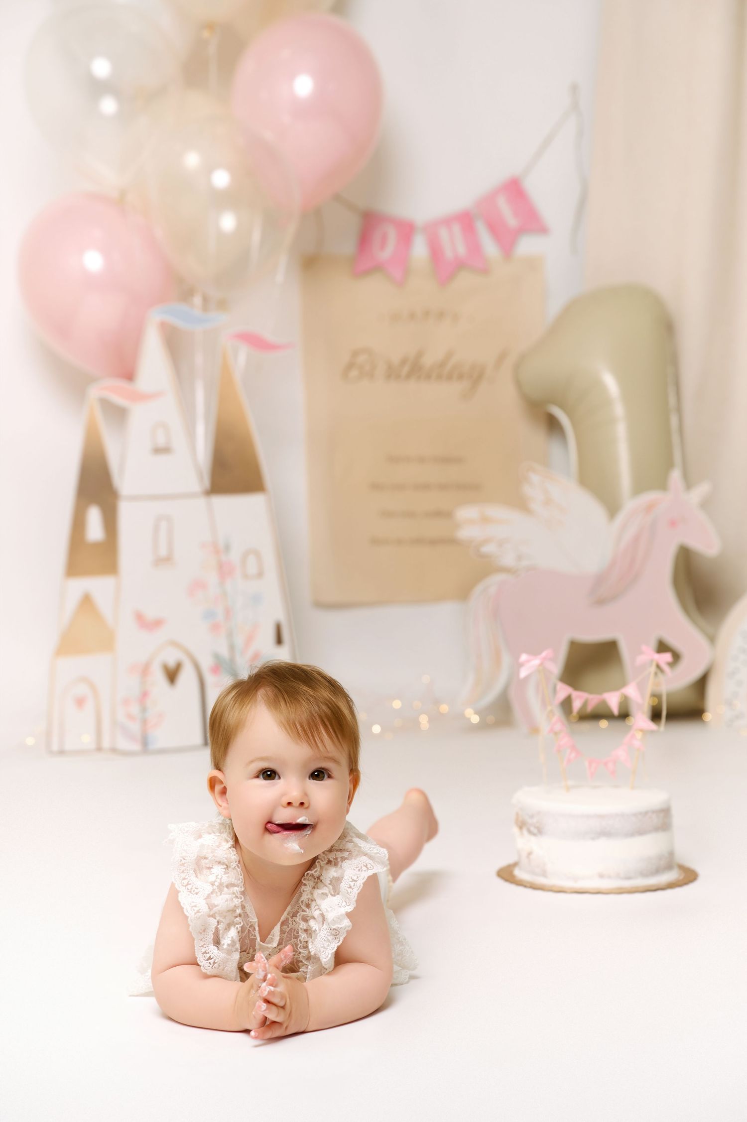 Baby girl celebrates first birthday lying on floor with unicorn cake, pink balloons, and castle decorations.