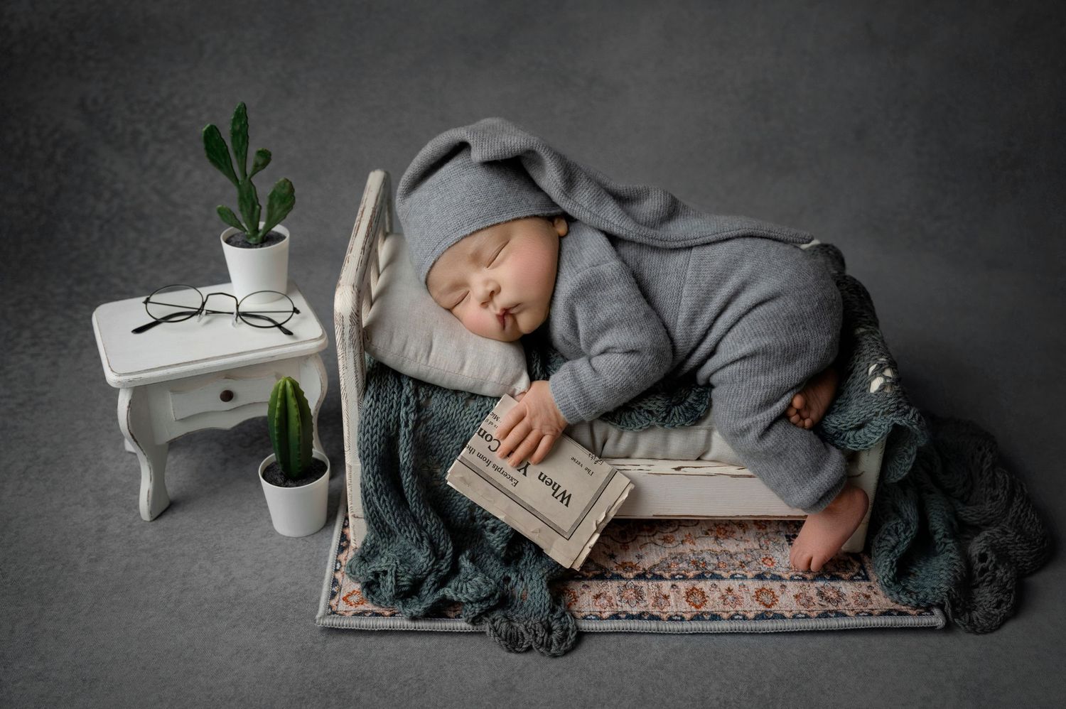 Newborn baby sleeping on tiny bed in gray knit outfit with bunny hat, holding book, beside small white table with glasses.