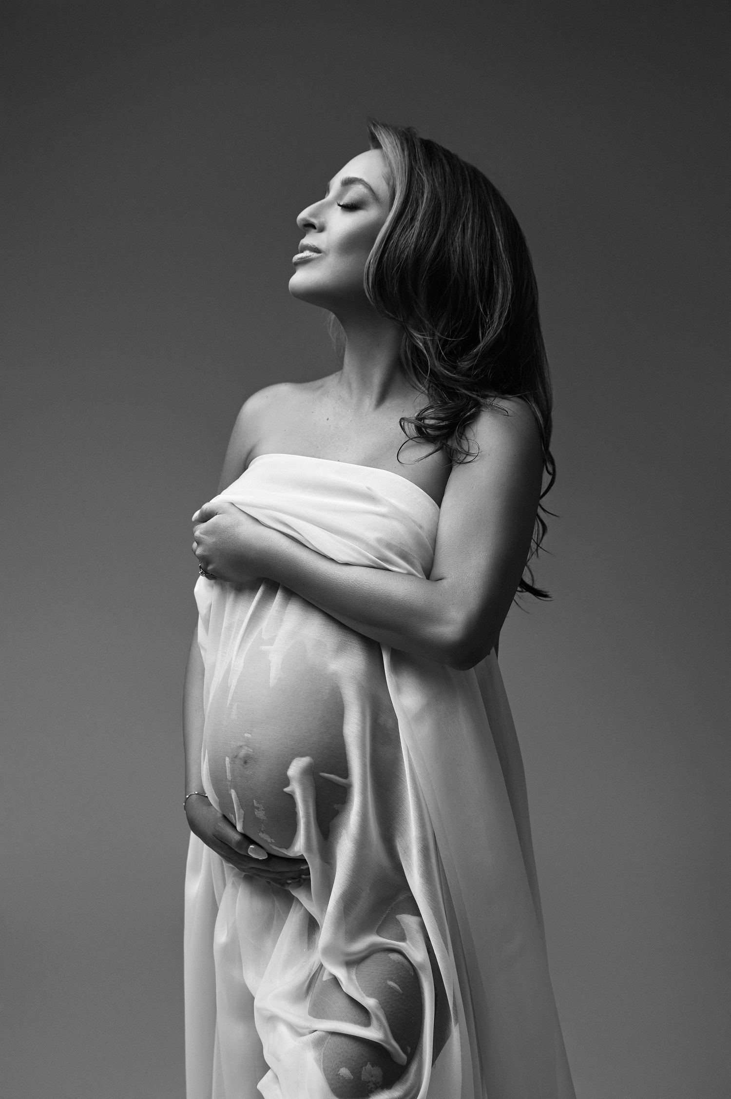 Black and white maternity photo of a pregnant woman draped in white fabric, cradling her belly with eyes closed.
