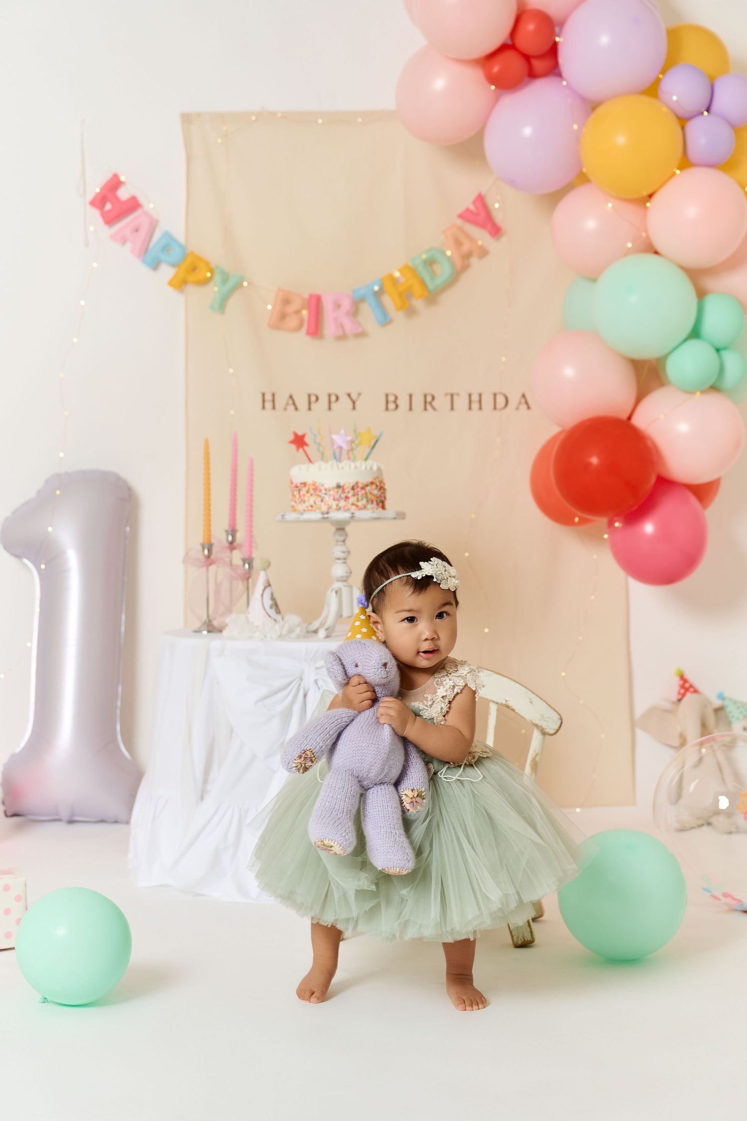 Adorable baby girl in green tutu dress holds stuffed bunny at colorful first birthday party with balloons and cake.