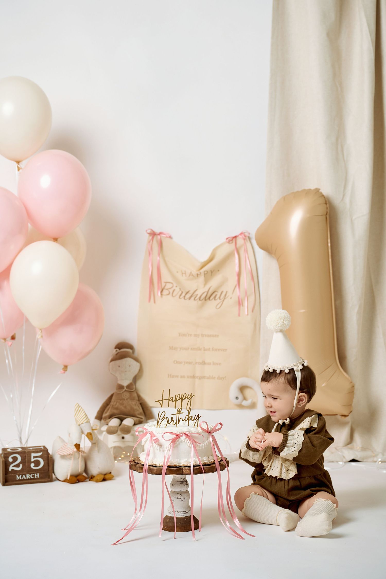 Baby girl celebrates first birthday with cake, pink balloons, and toys on March 25th.