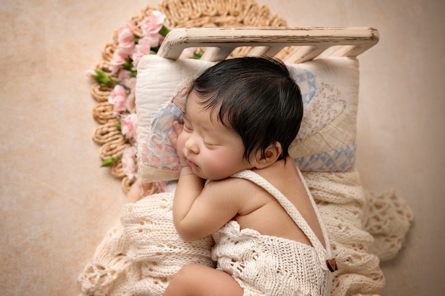 Newborn baby sleeping peacefully on a tiny bed with floral pillow, wearing a cream knit romper.