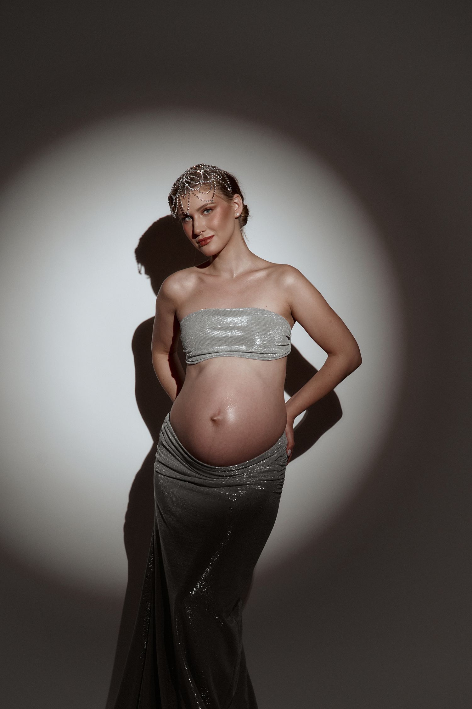 Pregnant woman in silver strapless crop top and skirt posing under spotlight, showing baby bump, studio maternity photo.