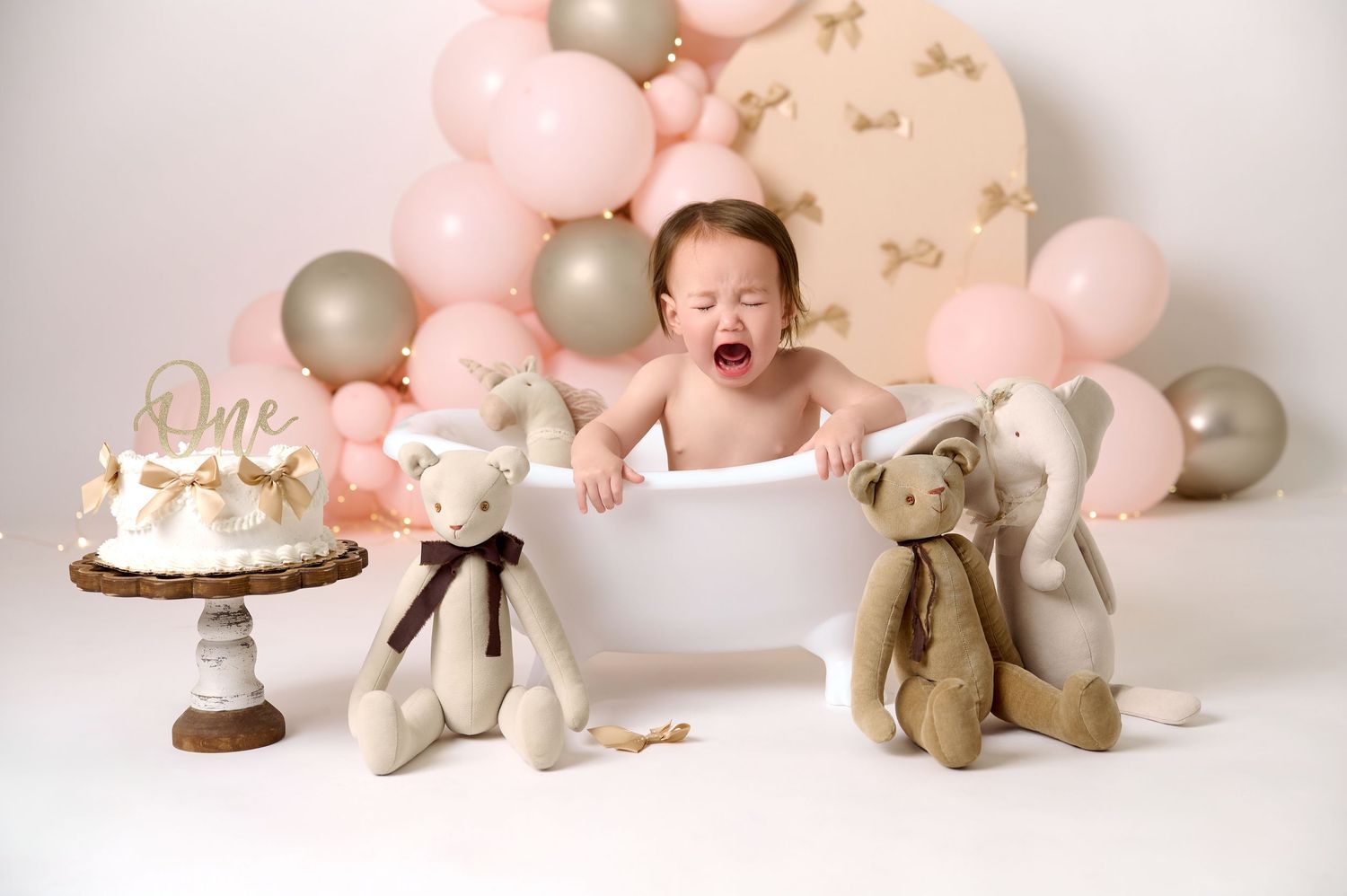 Crying baby in white tub at first birthday cake smash with pink balloons, stuffed animals, and a one cake topper.