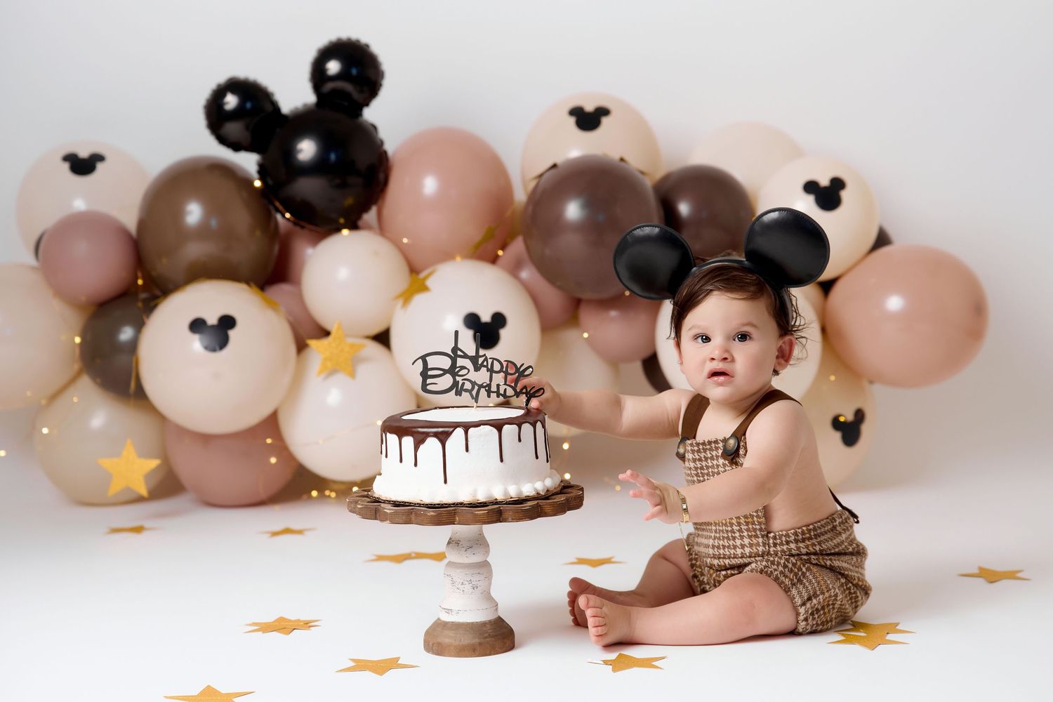 Baby's first birthday cake smash with Mickey Mouse balloons, plaid outfit, and chocolate cake on white background.